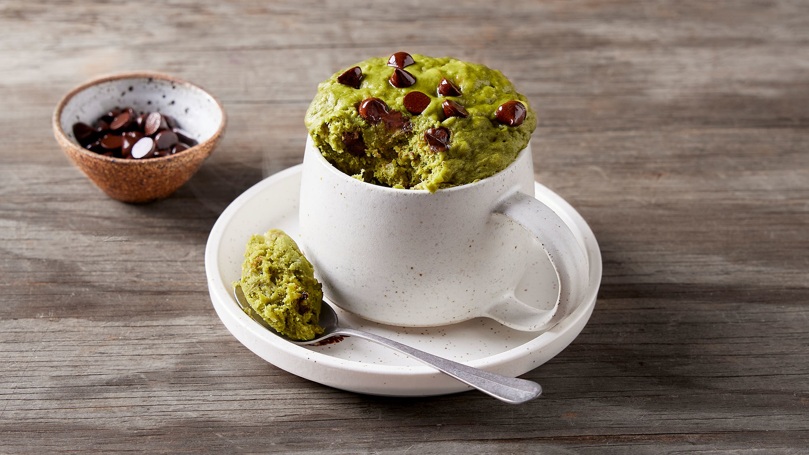 A white ceramic mug of luscious green Matcha, Banana & Choc Mug Cake, dotted with melting choc chips, sits on a matching plate, ready for eating.