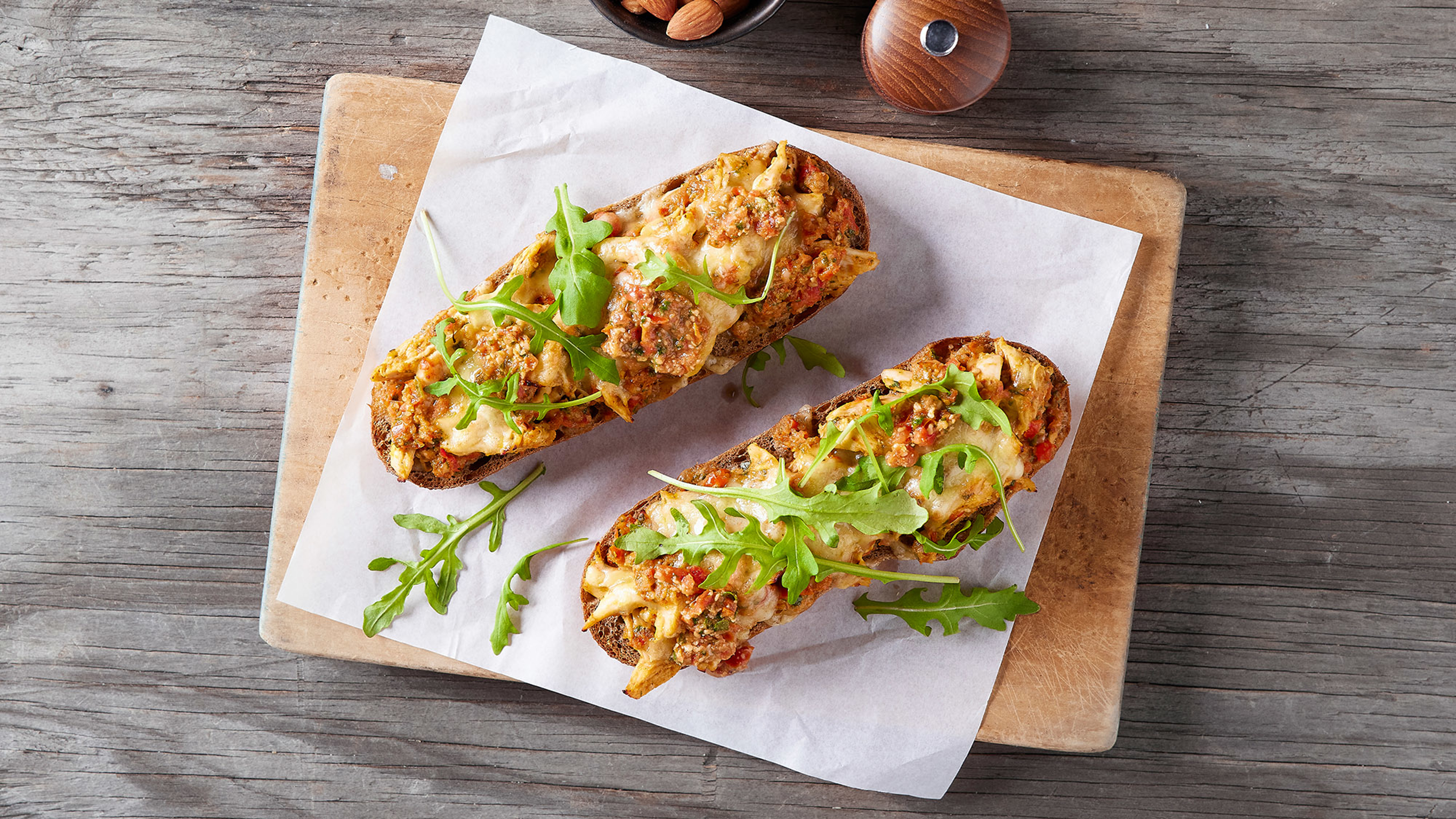 Two serves of Centr's Open Chicken & Pesto Sandwich sits on a square of baking paper, on a well-used wooden cutting board.