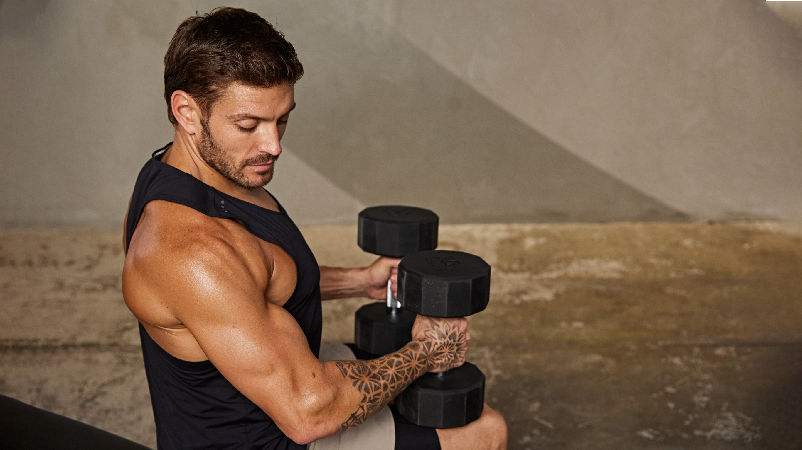 Luke Zocchi sits, holding 2 dumbbells on his thighs, looking at his flexed, muscular right arm.