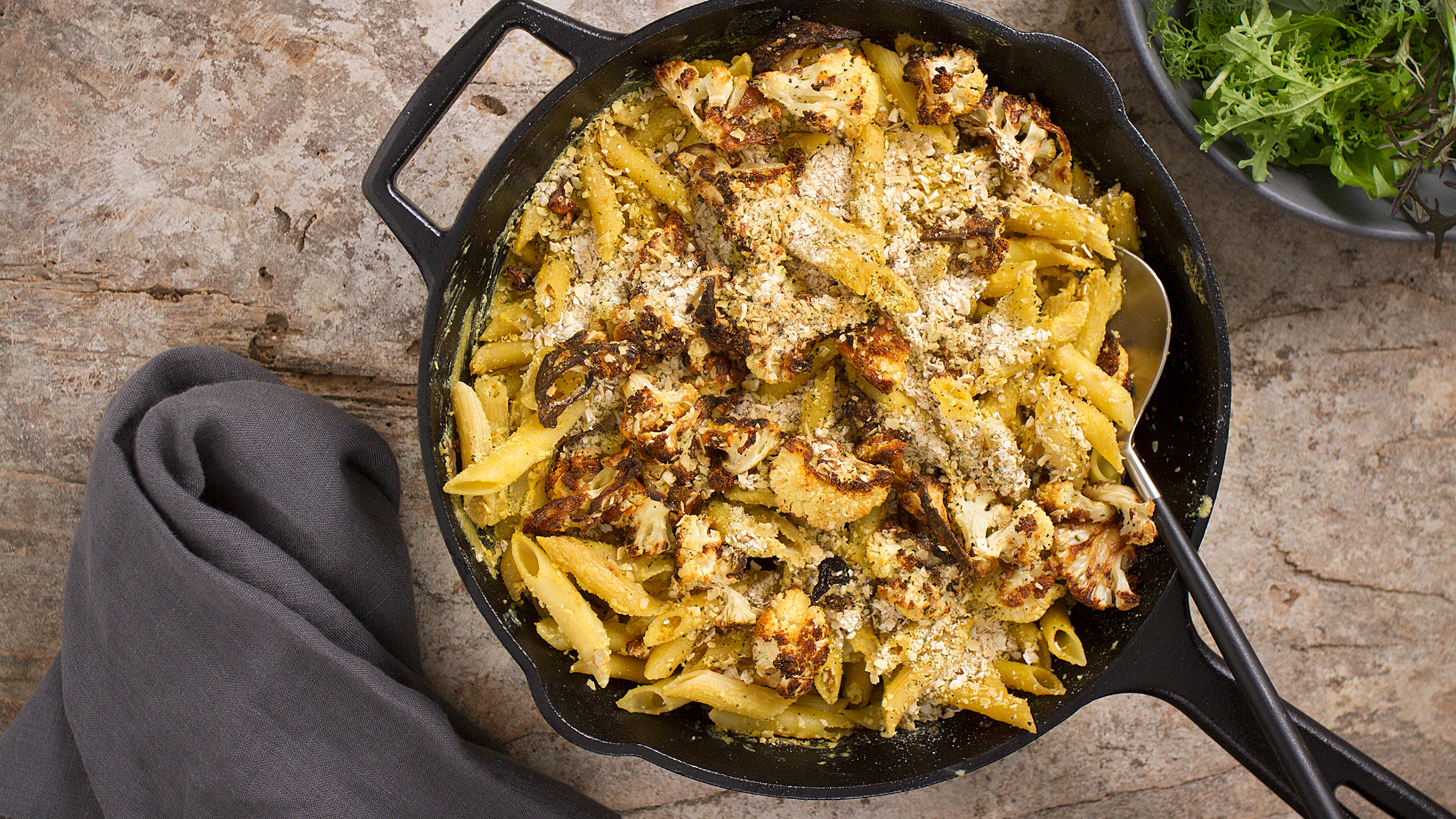 A skillet pan of Dan’s Vegan Cauliflower Mac & Cheese rests on a board, ready for serviing.