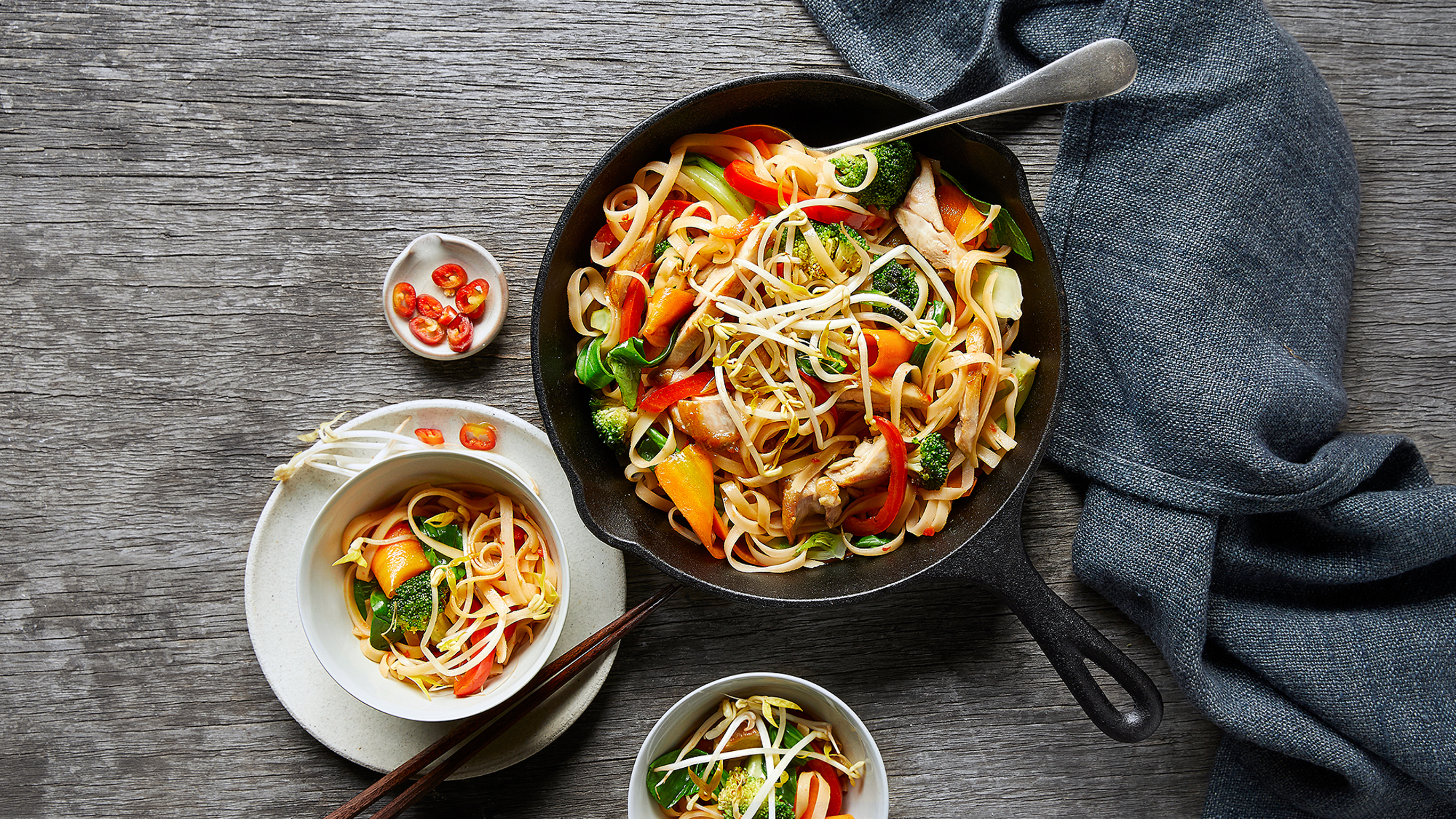 A freshly plated erve of Centr's Chinese Chicken Stir-fry with Noodles sits beside a cast iron wok of Chinese stir-fry and noodles.