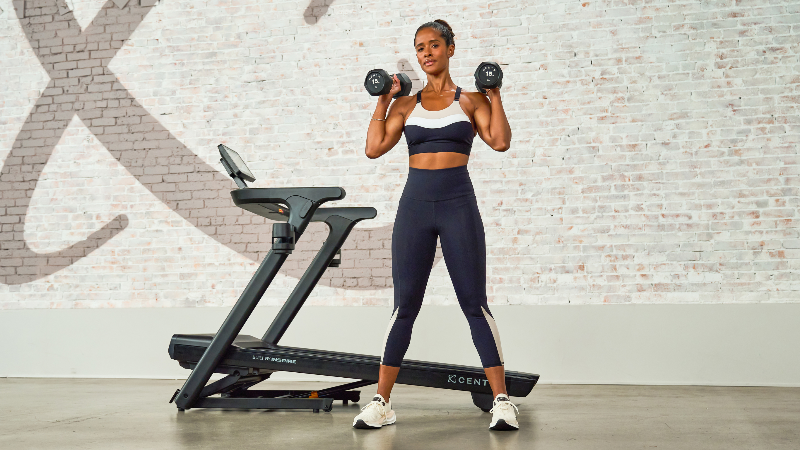 Centr trainer Ingrid Clay poses in front of the Centr's treadmill, with 2 dumbbells racked on her shoulders.