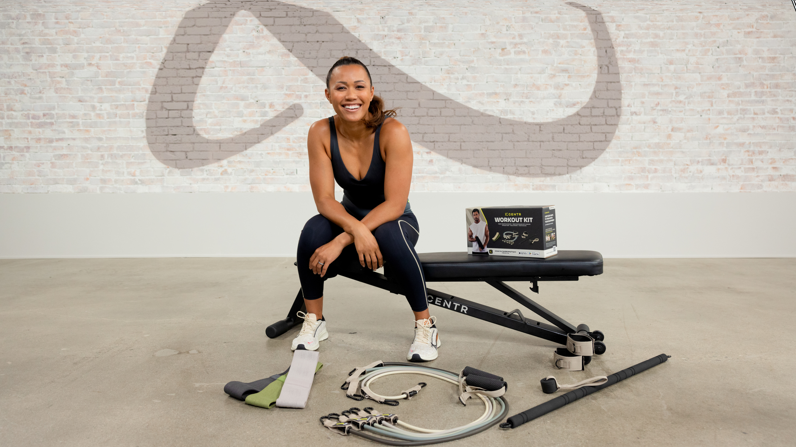 Centr trainer Maricrix Lapaix, sits on a gym bench in the Centr studio gym, surrounded by pieces of the Centr fitness kit.