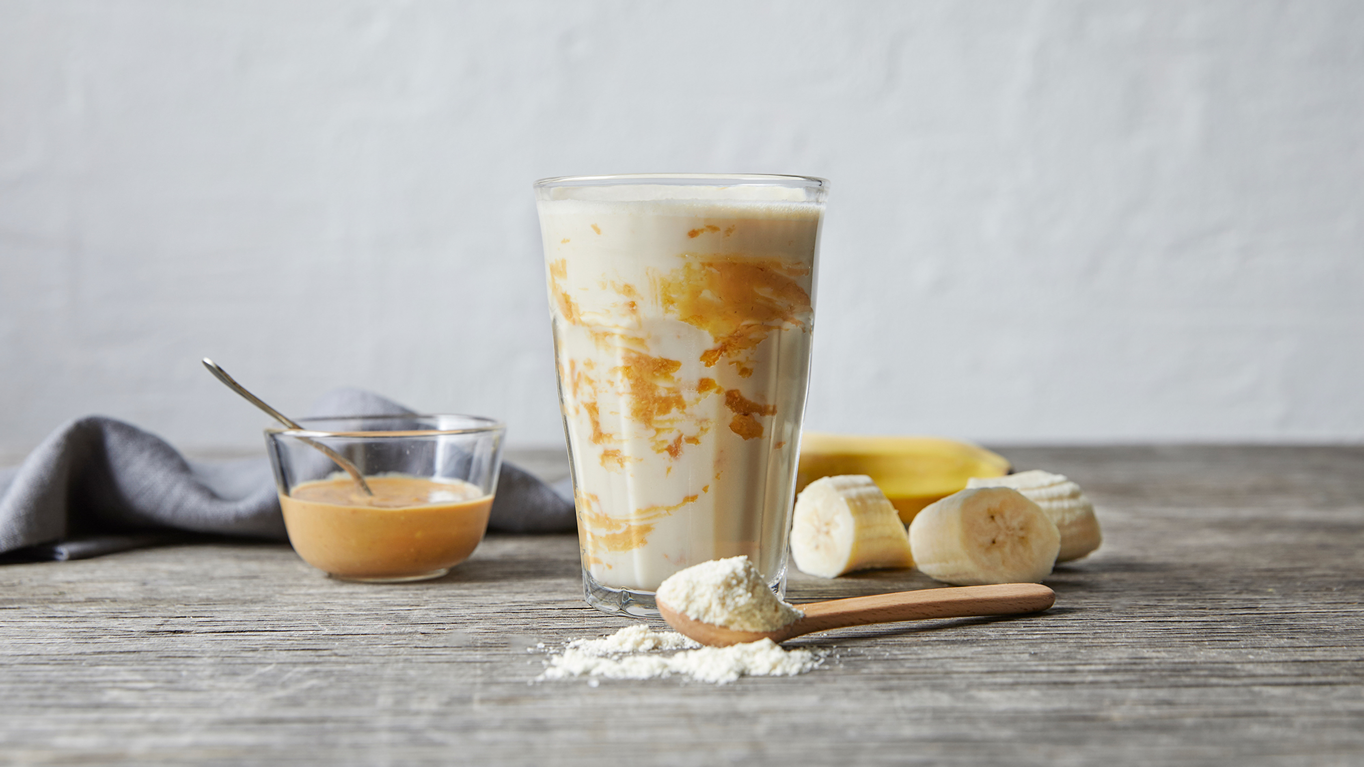 A glass of Centr's Banana Peanut Butter Snack Smoothie rests on a grey rustic wooden table, surrounded by thick sices of banana.