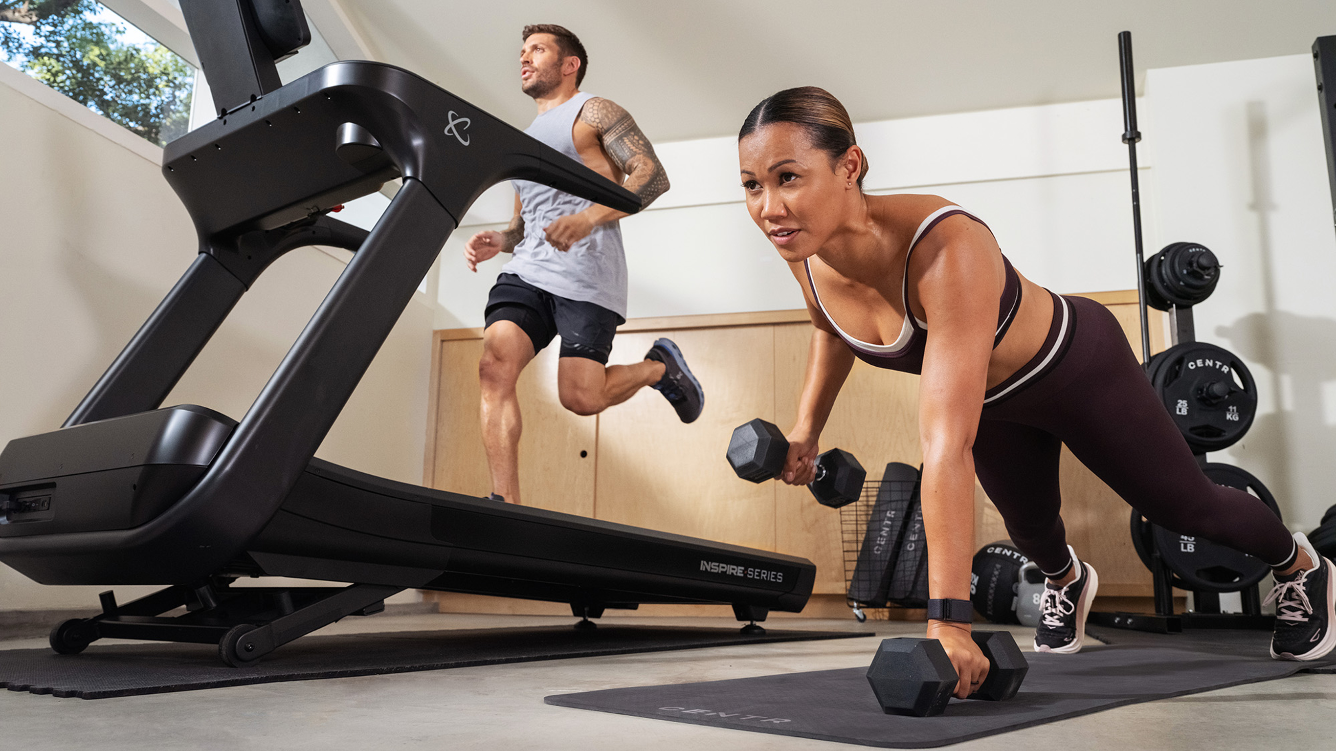Low angle photo of Centr trainer Maricris Lapaix performing dumbbell renegade rows on the floor beside the treadmill Centr trainer Luke Zocchi is using.