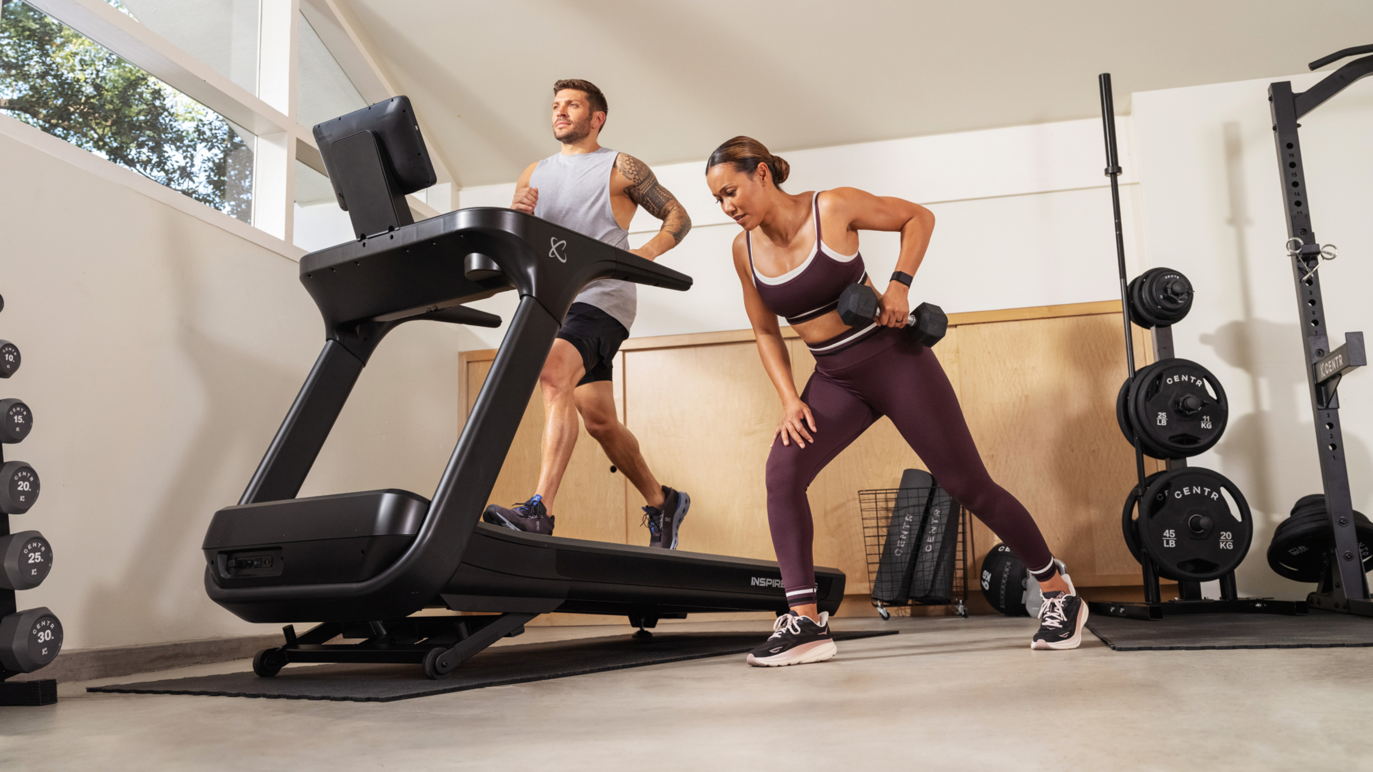 Centr trainer Luke ZOcchi works out on a treadmill while Maricris Lapaix performs dumbbell rows alongside the treadmill.