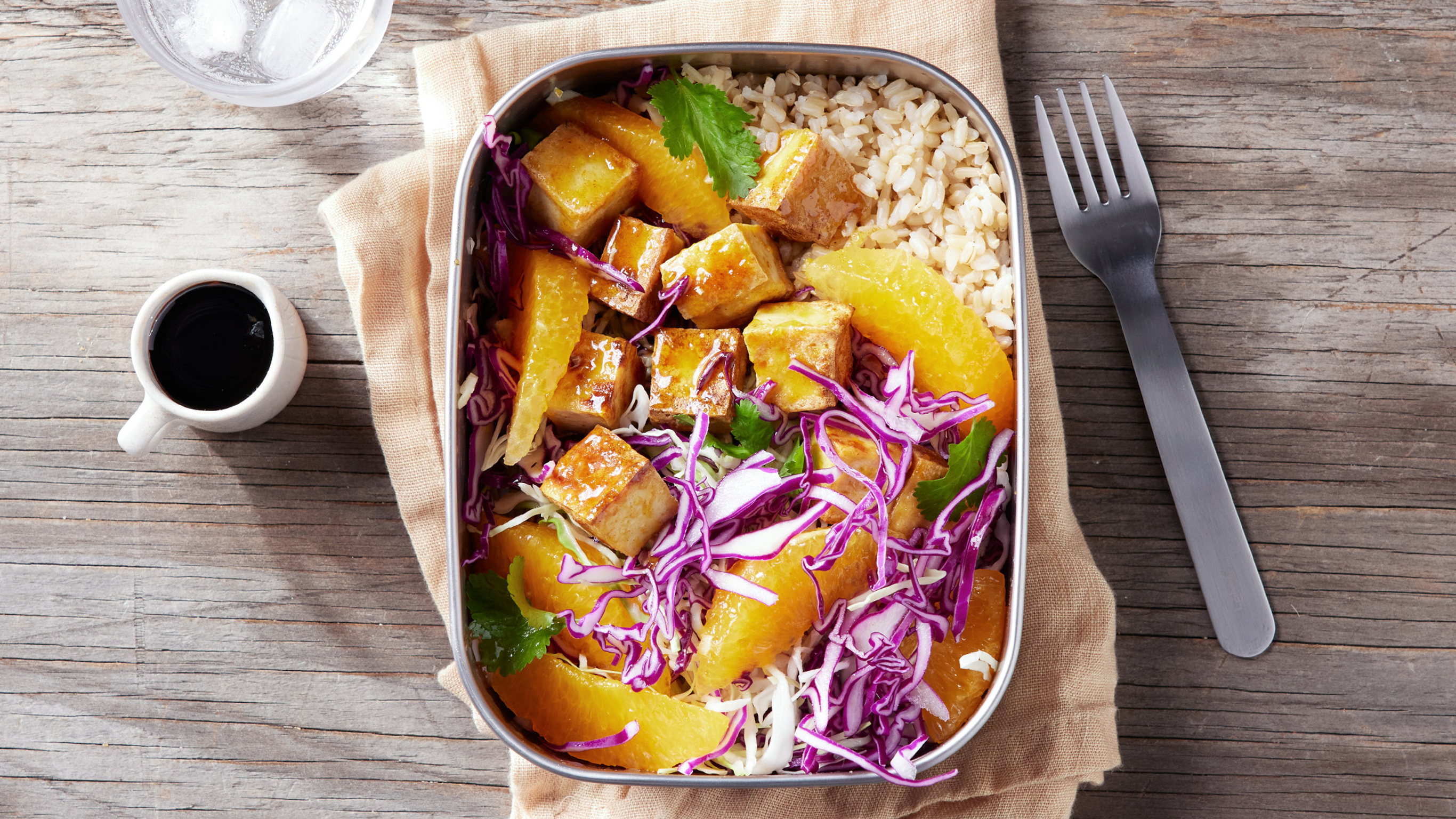 A metal llunch box of Centr's Orange Tofu & Brown Rice Bento Box sits on a linen napkin, fork on the side, ready for eating