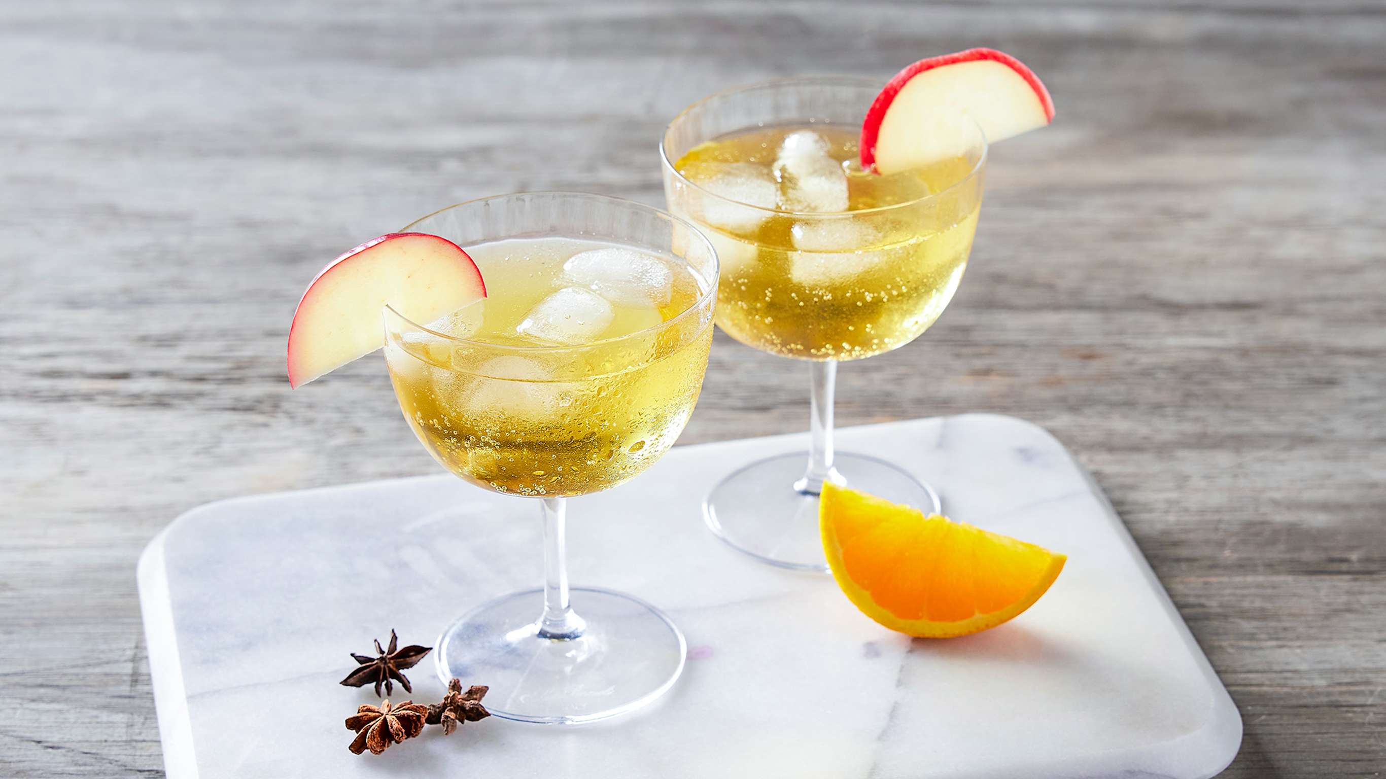 Two wine glasses of Centr's Spiced Apple Cider Spritz stand on a pale marble cutting board, ready for drinking.