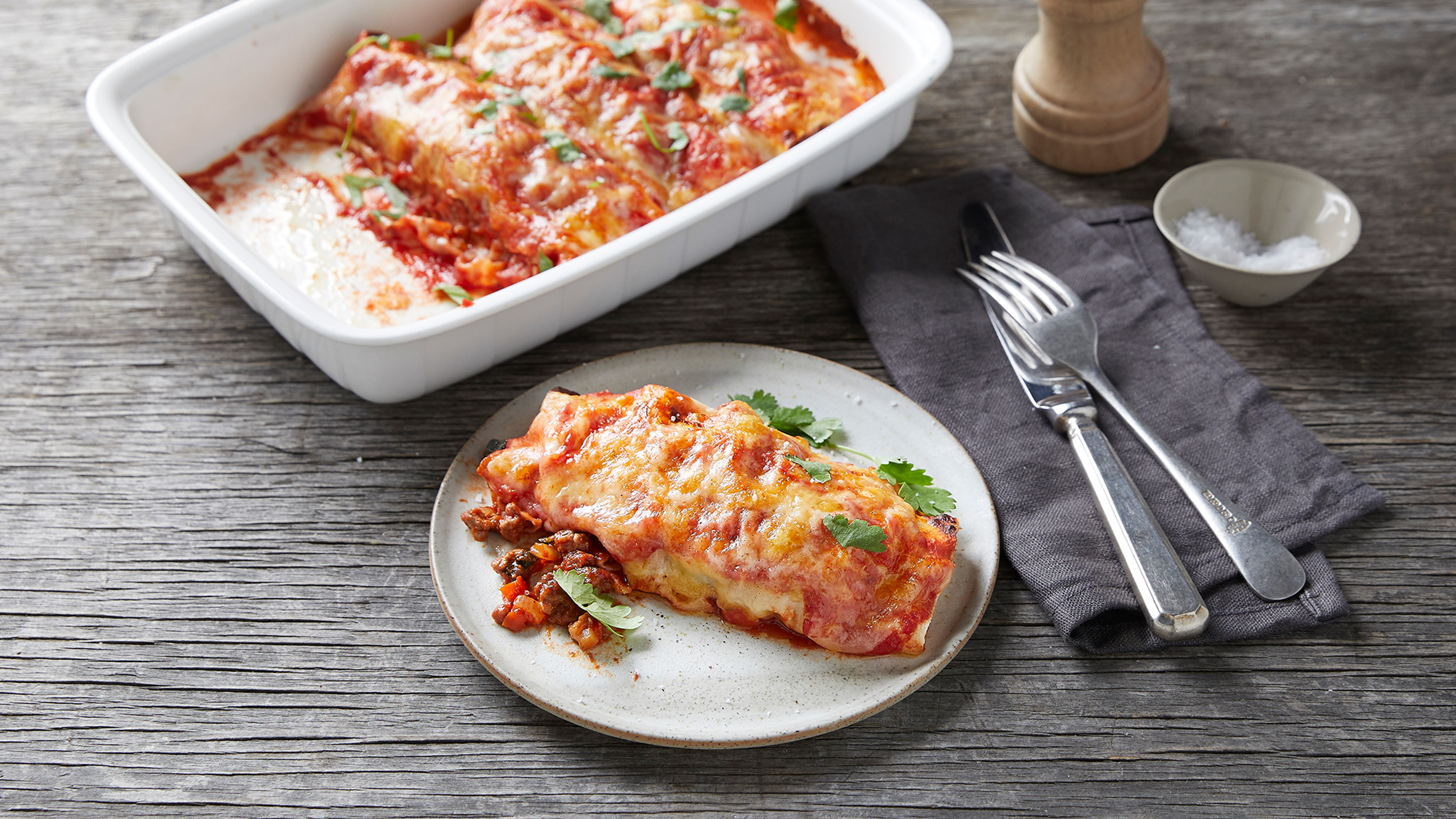 A serve of Centr's Beef and vegetable enchilida sits on a plate beside a baking tray of enchilidas