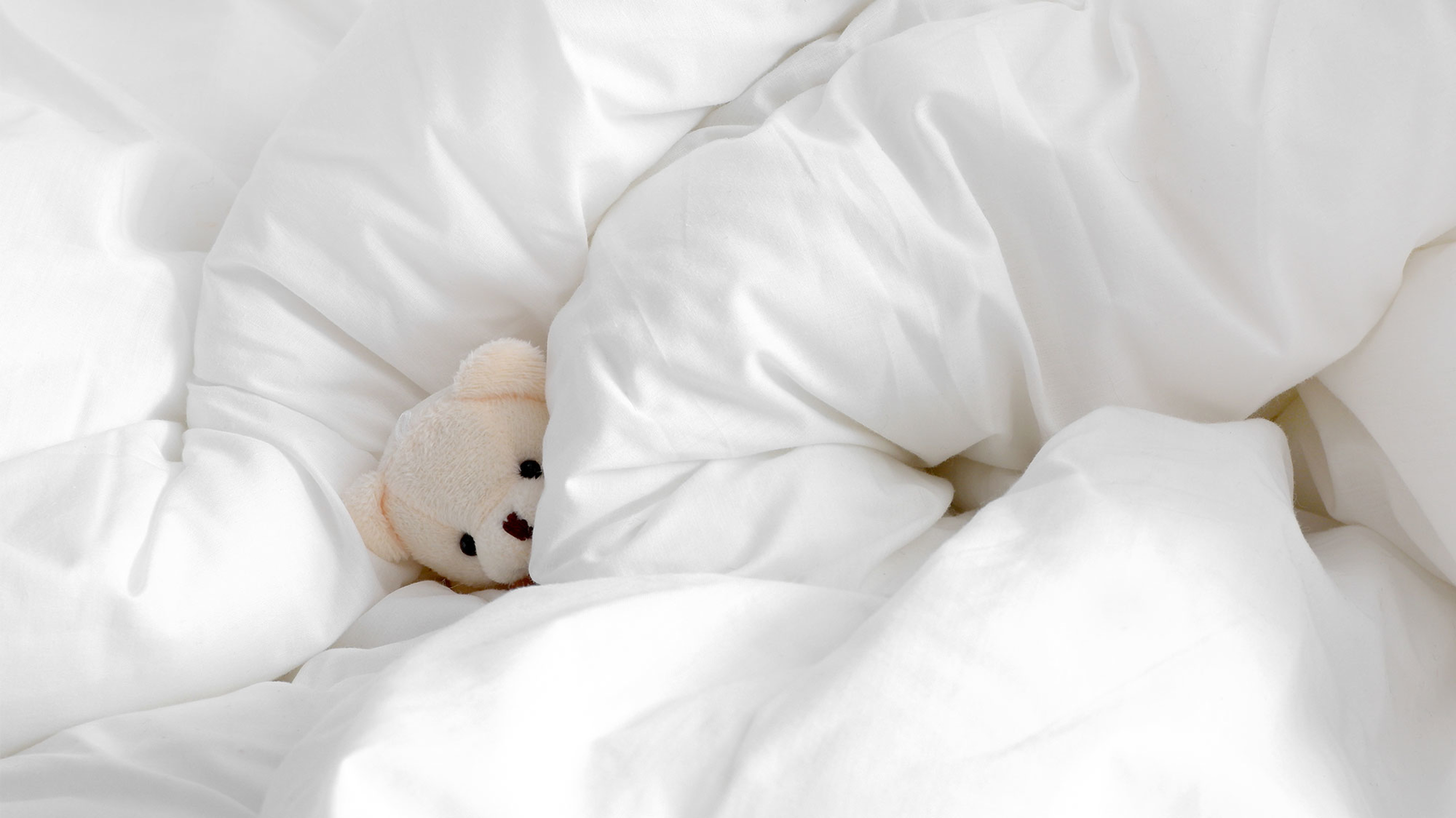 A photo a small white teddy bear tucked into a fluffy white doona and pillow.