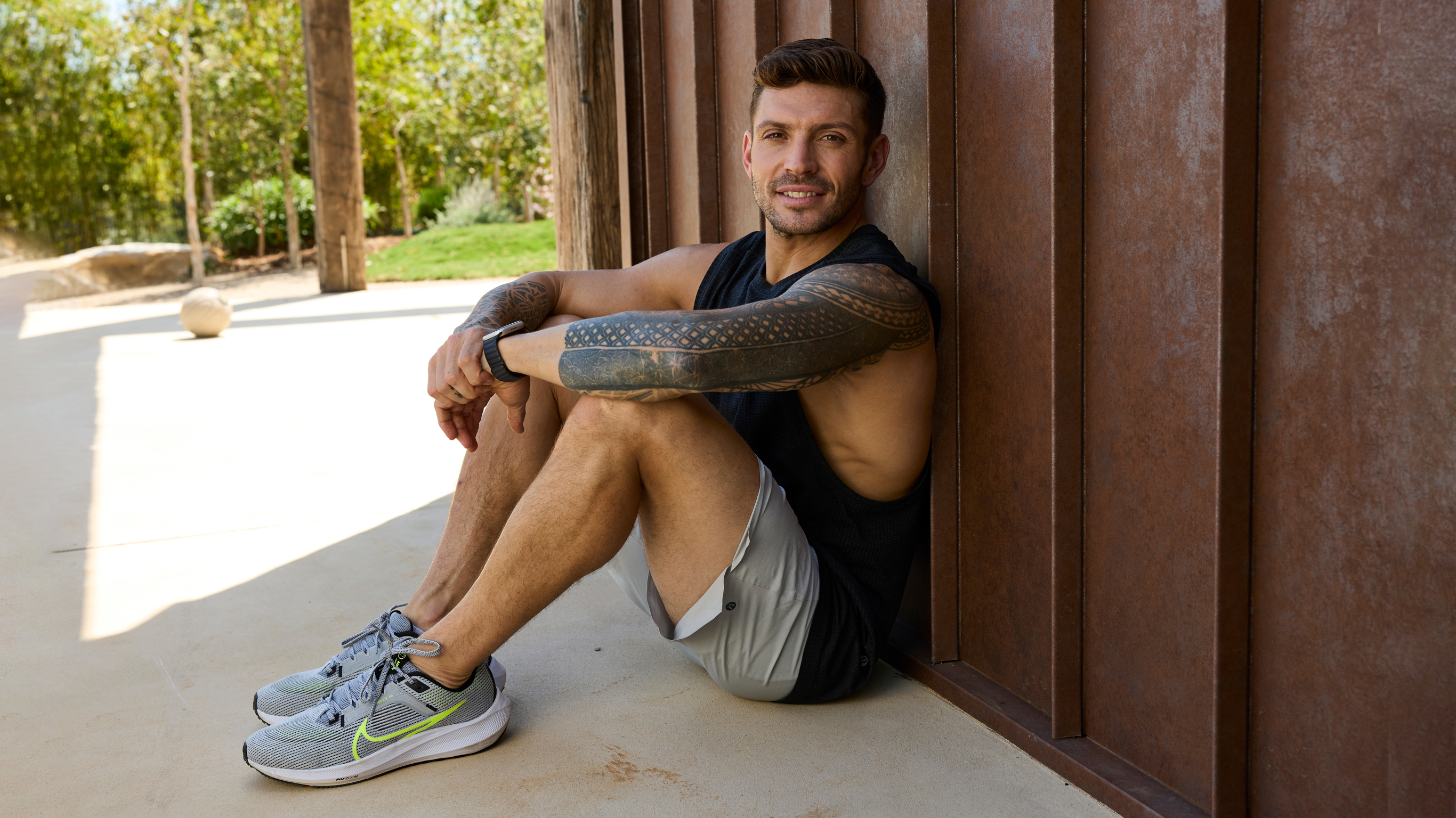 Centr trainer Luke Zocchi sits on the ground, leaning back against a rust-coloured metal wall, with his arms resting on his raised knees.