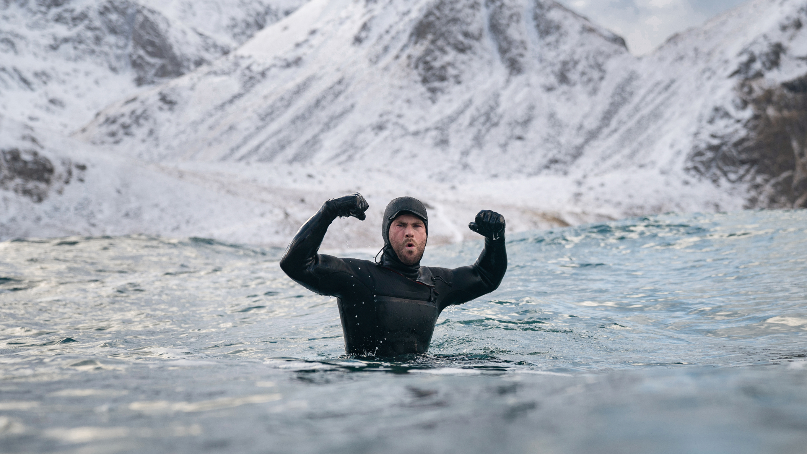 A photo of Chris Hemsworth in a full body wetsuit, arms raised and flexed, in churning icy ocean waters.