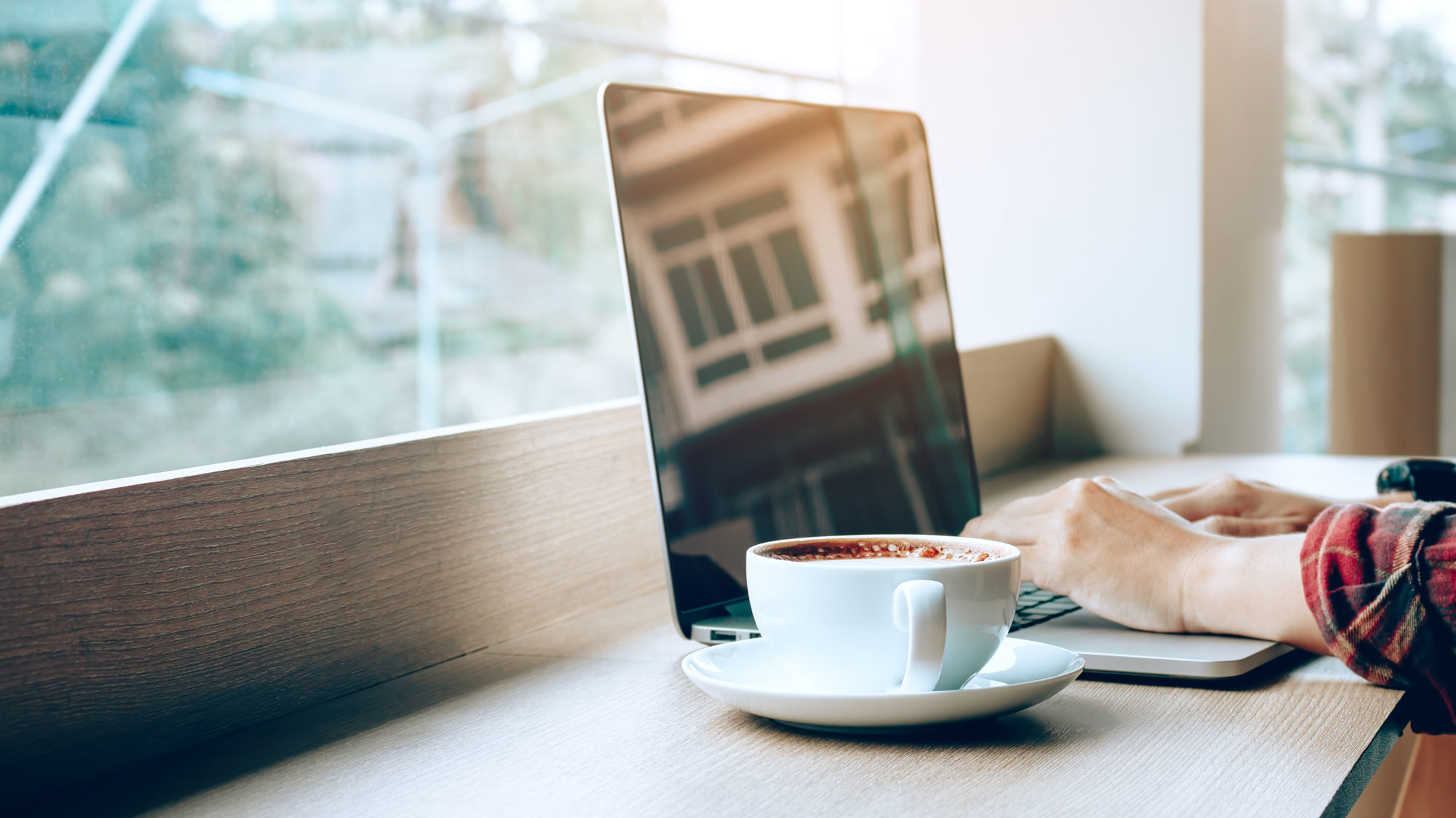 A photo of a white cup of coffee beside an open laptop that someone  is working at