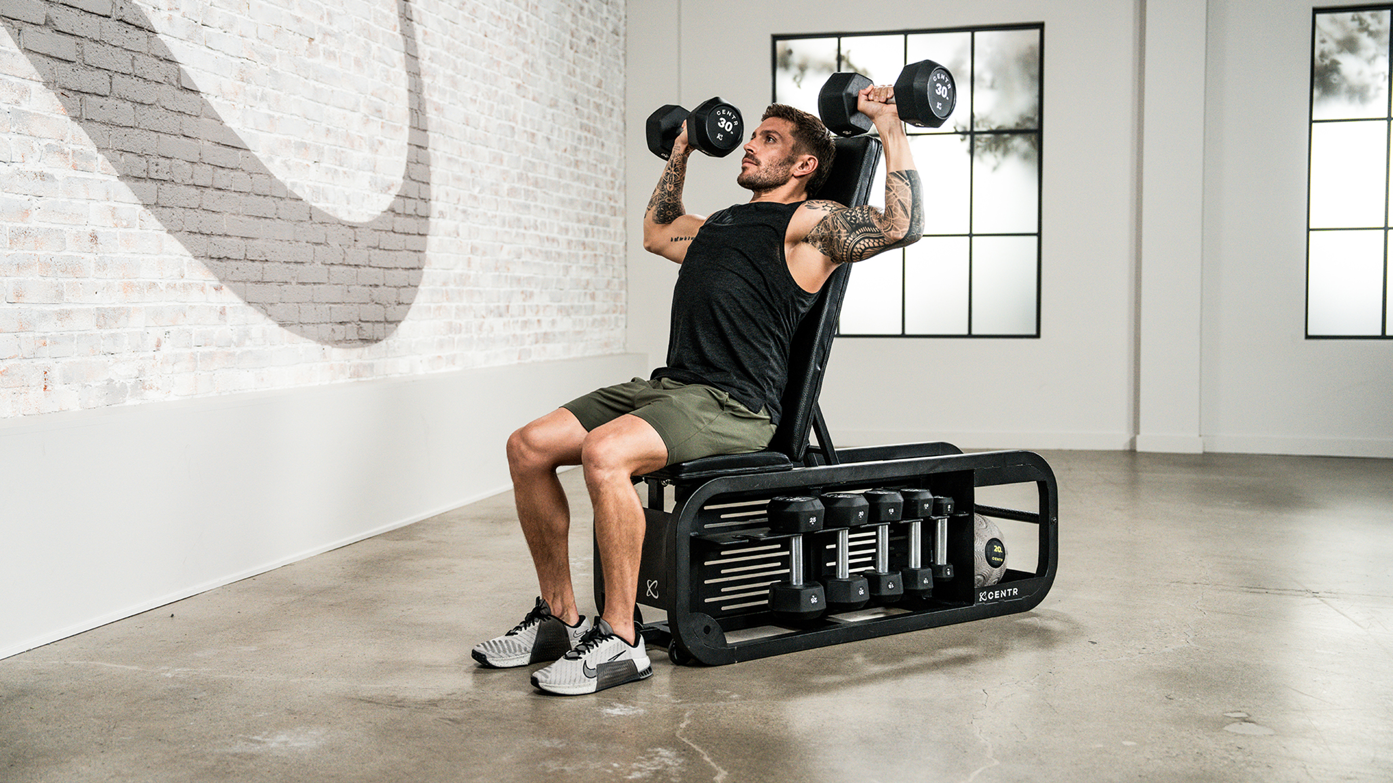 Centr trainer Luke Zocchi. performs dumbbell shoulder press while seated on the end of a Centr Fusion bench