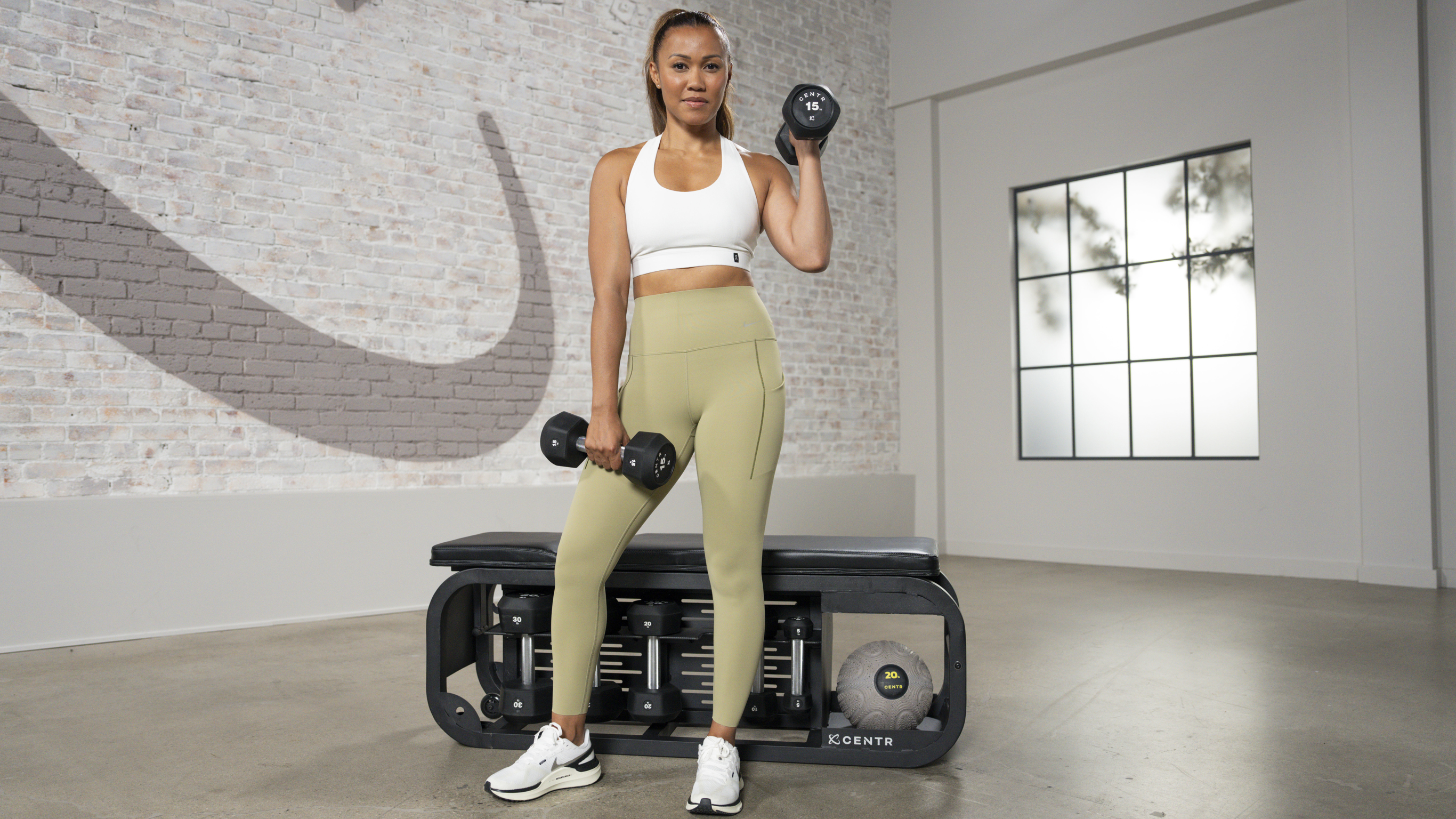 Centr trainer Maricrix Lapaix, poses with dumbbells in front of a workout bench, while looking at the camera.