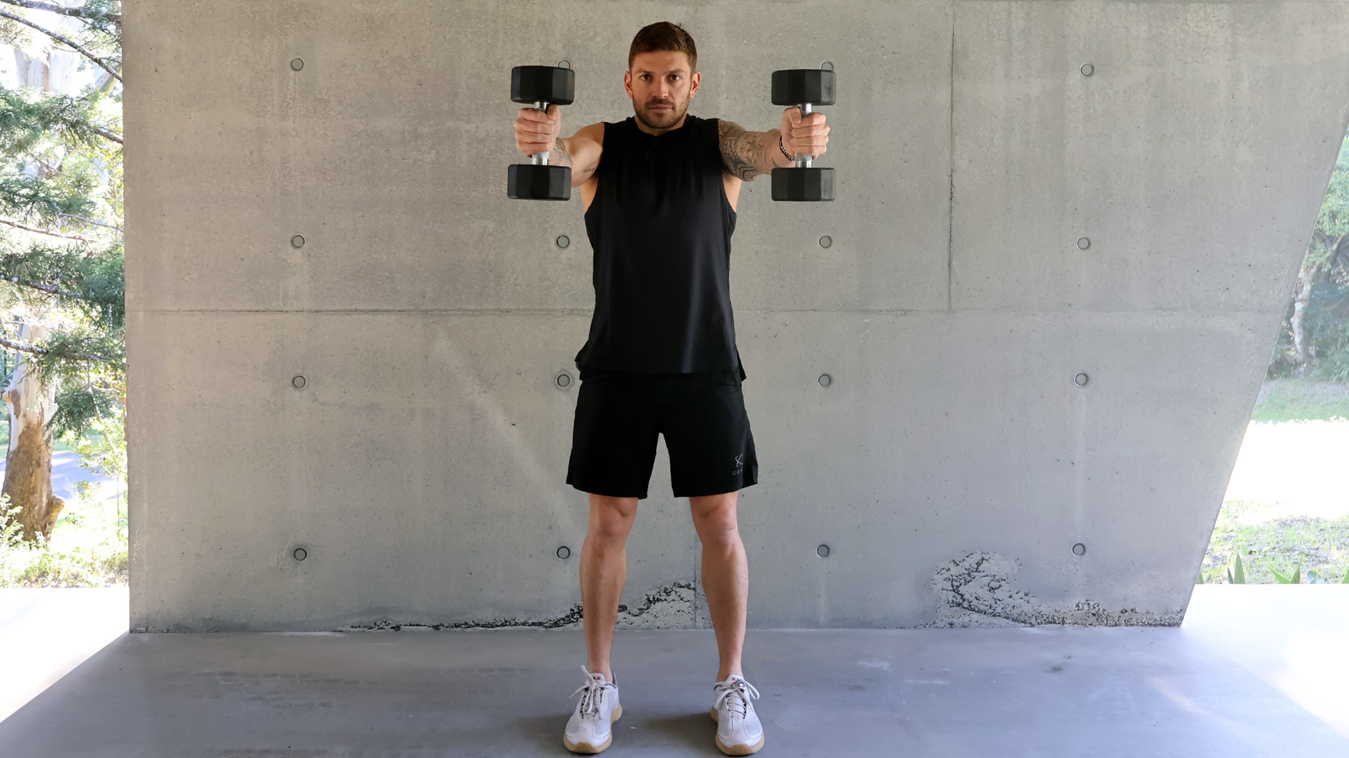 Luke Zocchi holds a pair of dumbbells directly out in front while looking at the camera