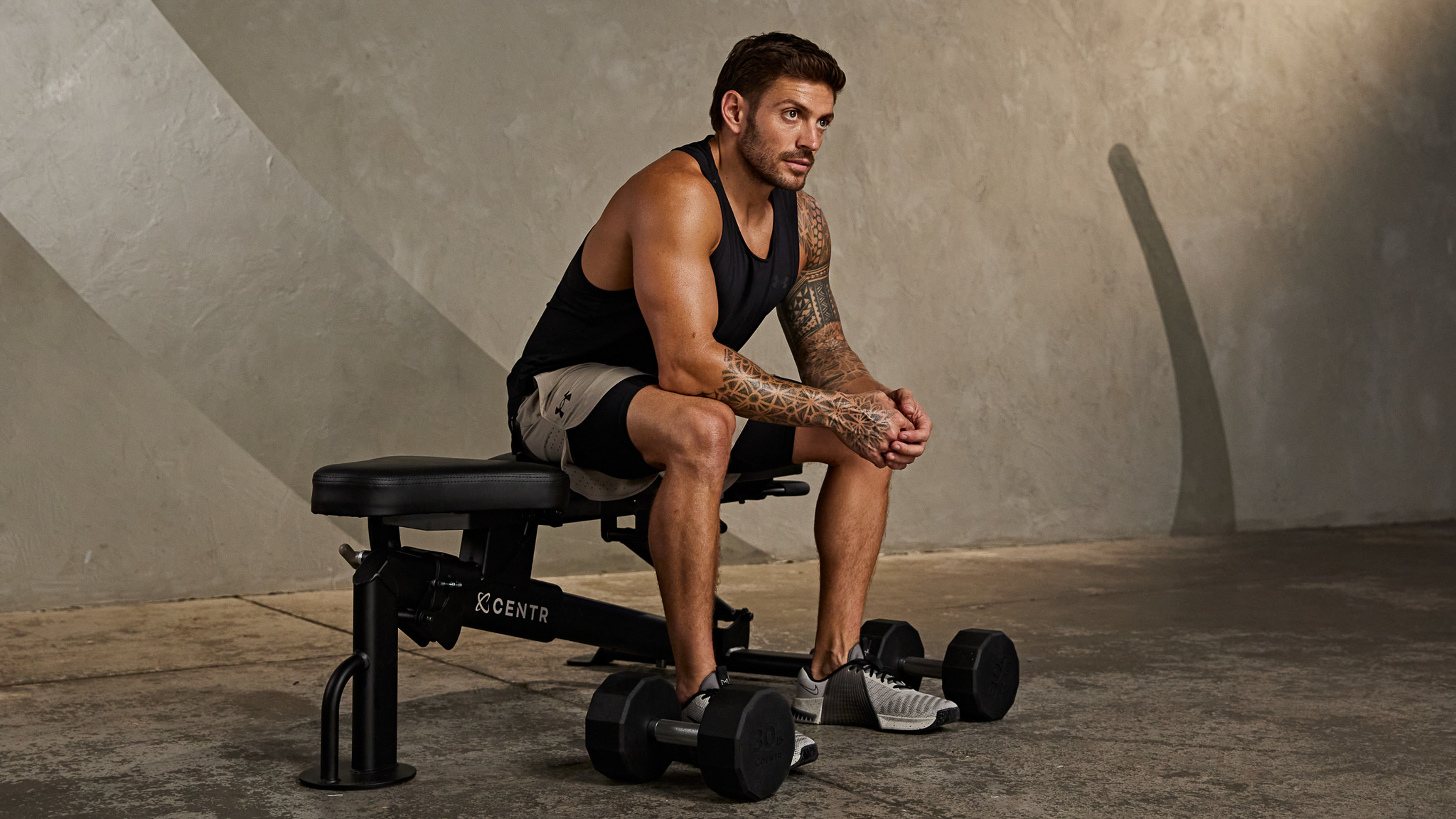 Centr trainer Luke Zocchi sits on a workout bench, elbows on knees, looking out, with a pair of dumbbells at his feet.