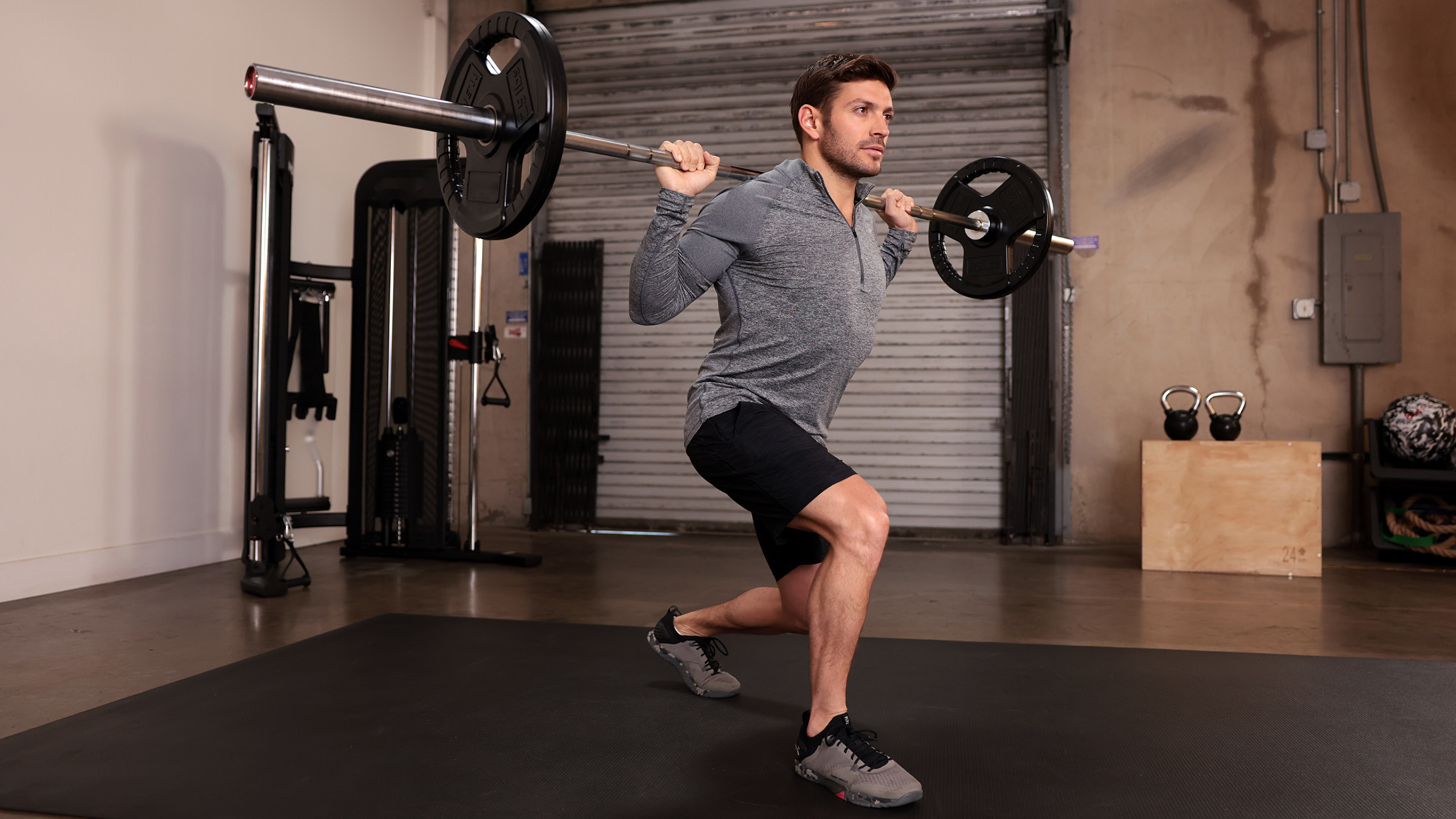 Centr trainer Luke Zocchi performs a forward lunge with weight plates on an olympic bar in a garage gym.