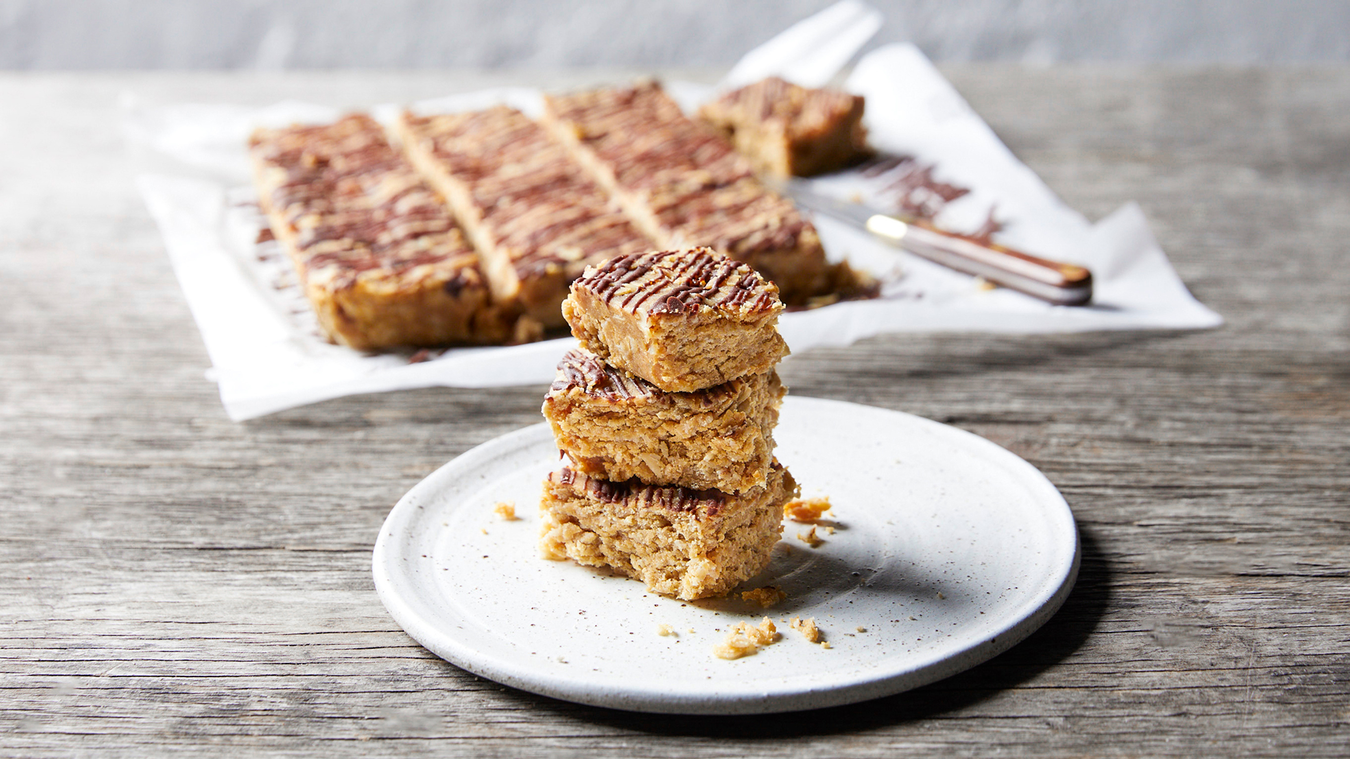 Three square pieces of Centr's Peanut Butter & Honey Protein Squares are stacked in a tower on a white round plate.