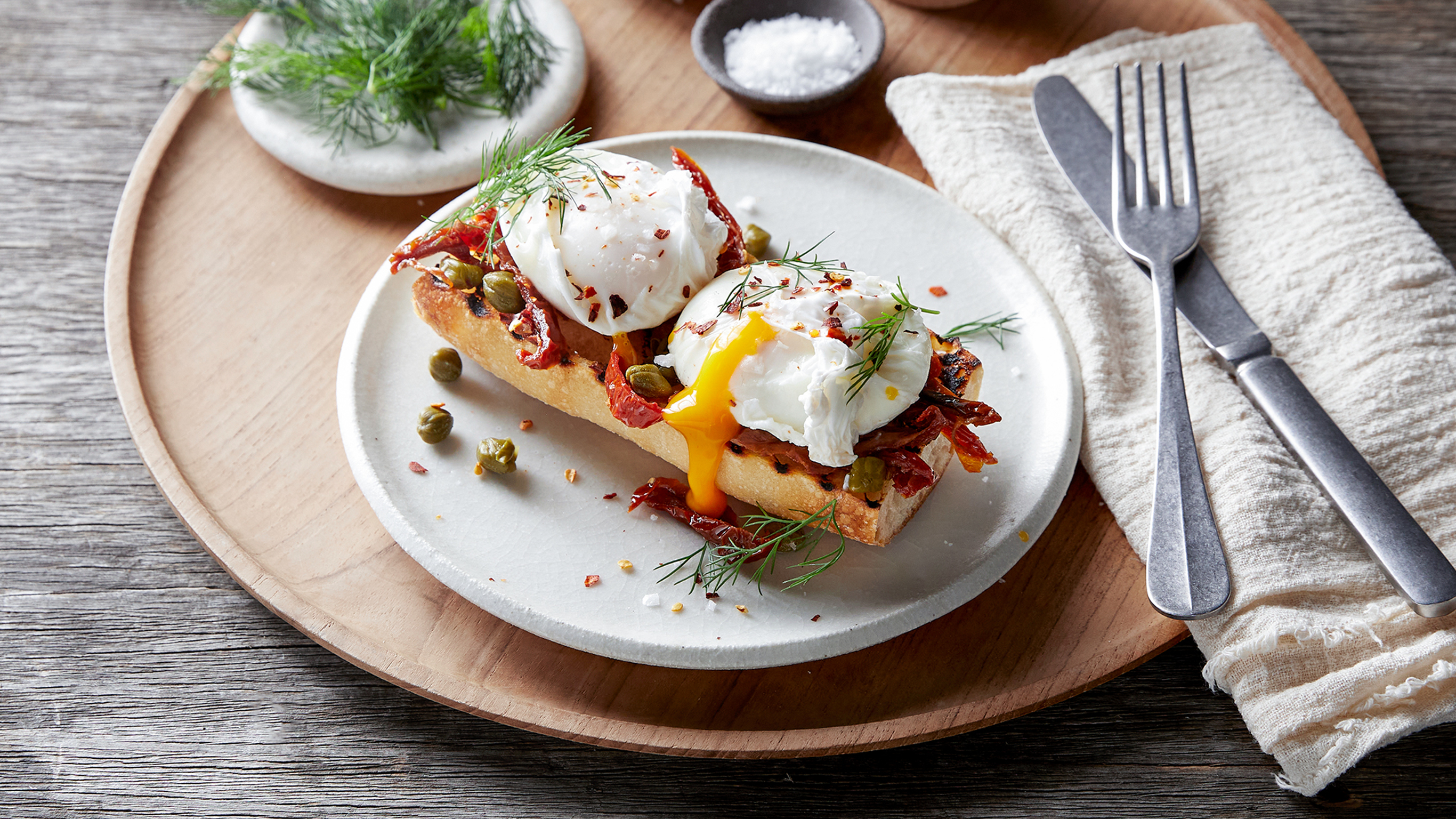 A serving of Poached Eggs on Toast with Capers & Sundried Tomatoes sits on a round white plate, ready for eating.