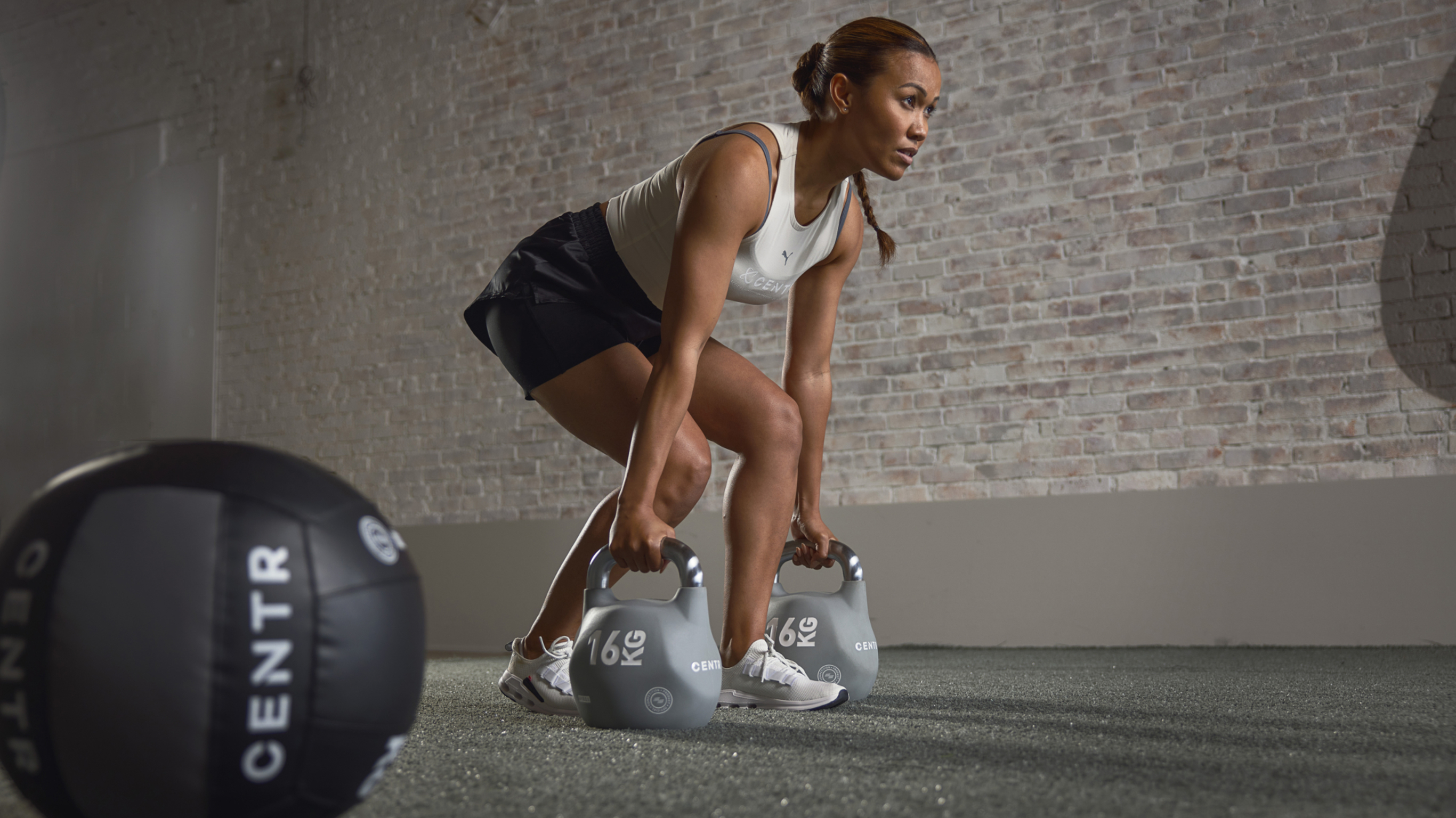 Maricrix Lapaix bends over to pick up a pair of Centr kettlebells in a split stance RDL