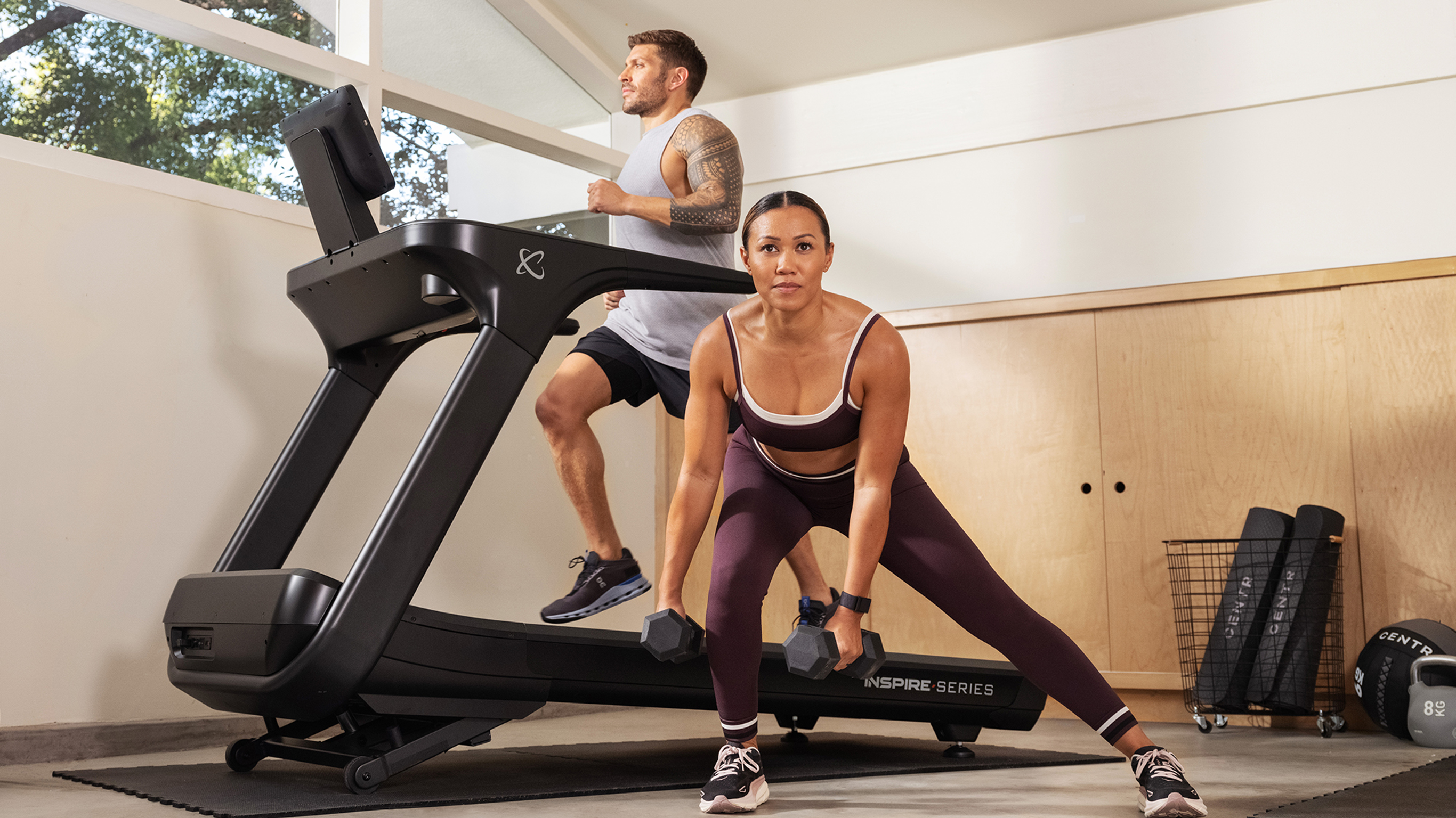 Luke Zocchi runs on a Centr treadmill while in front, of the treadmill, Maricris Lapaix performs a side lunge with Centr dumbbells.
