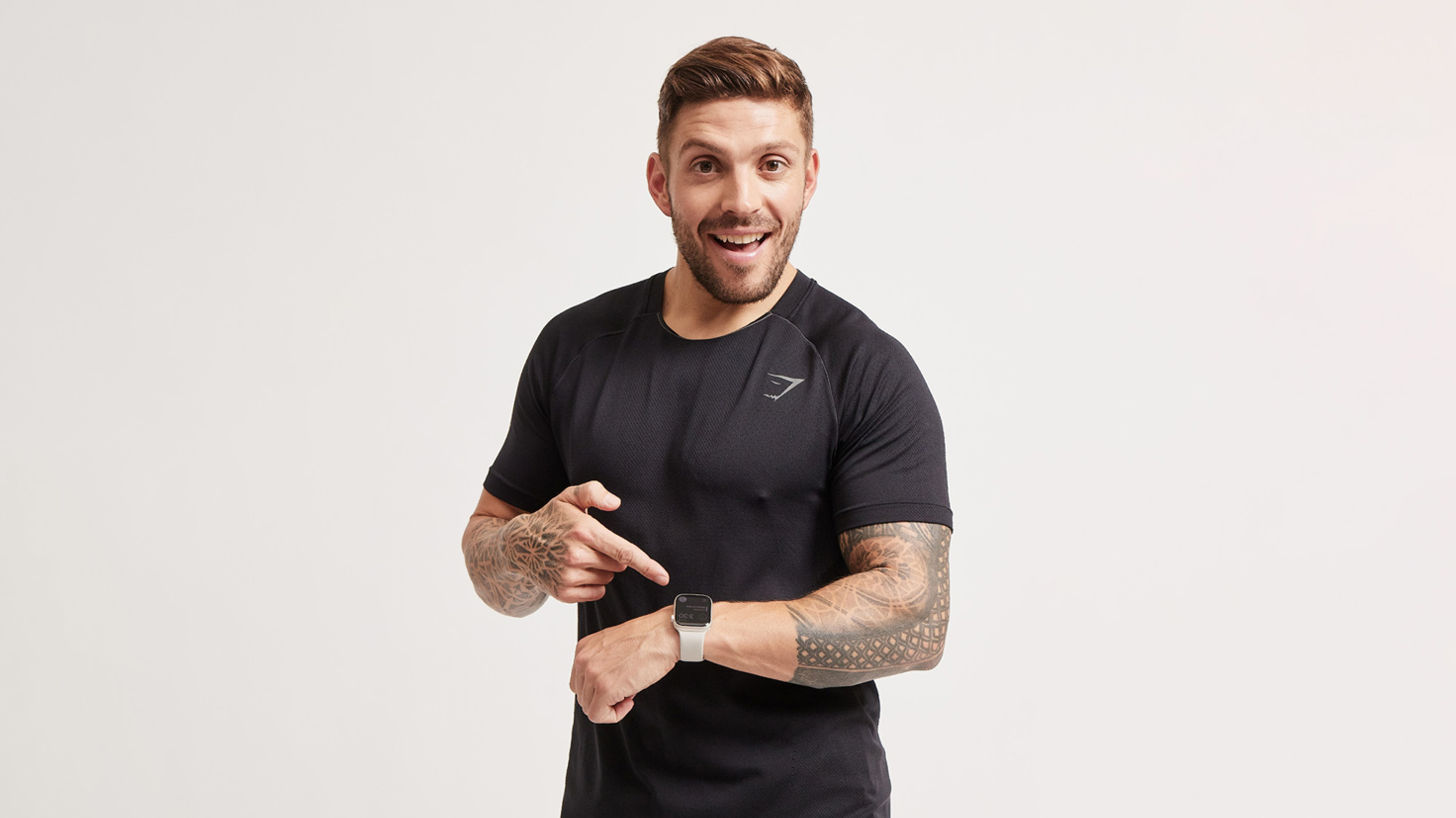 Luke Zocchi points to his smart watch on his wrist while looking at the camera with an open mouth, in amazement.