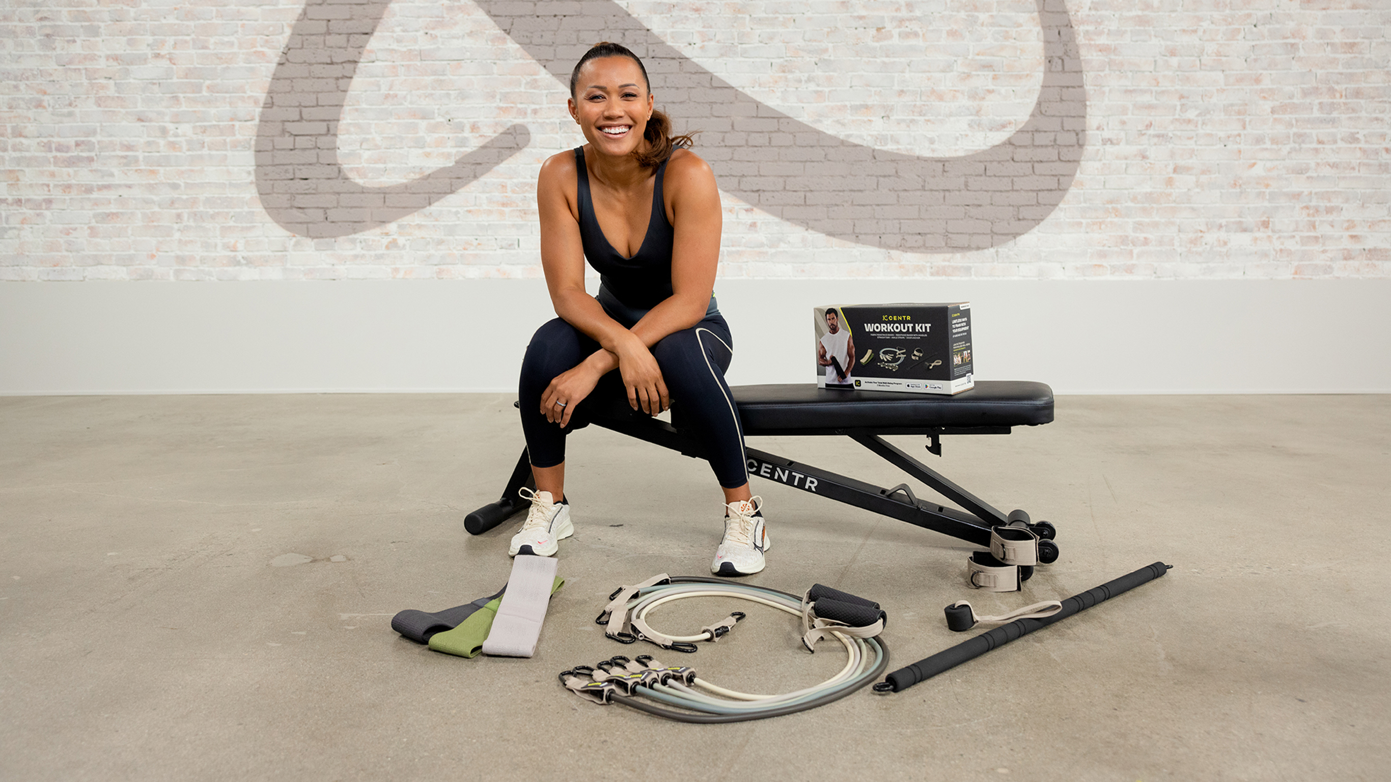 Maricris Lapaix sits on a workout bench with the contents of the Centr workout kit laid out around her on the floor.