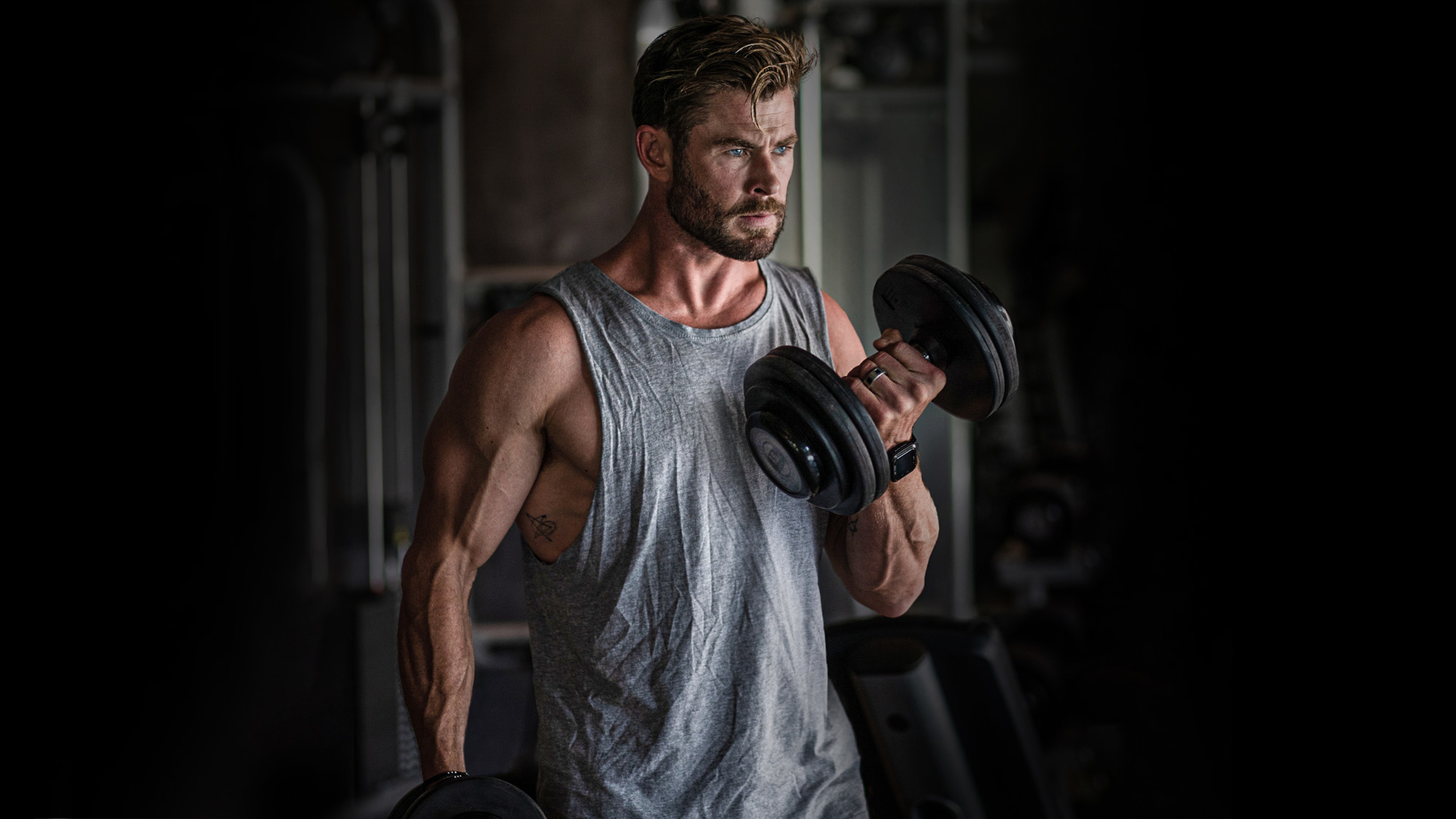 Chris Hemsworth, wearing a singlet top in a darkened gym, performs bicep curls with dumbbells.