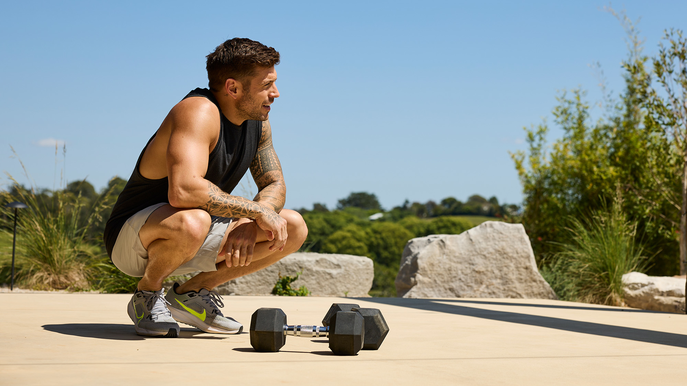 Luke Zocchi  squats, elbows on knees, resting between sets,  with a pair of Centr's hex dumbbells at hs feet