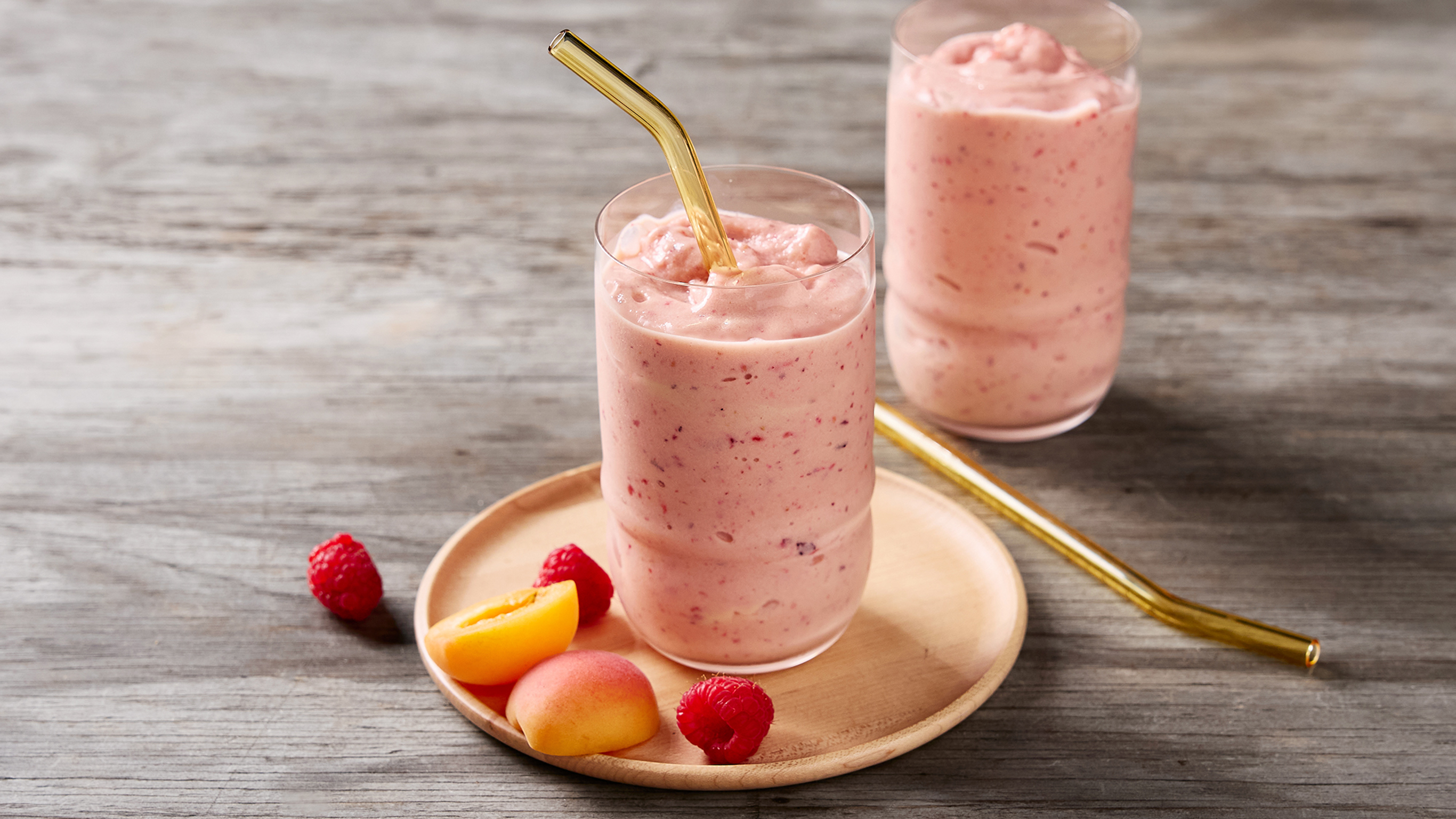 Two tall glasses, one a plate surrounded by pieces of peach, of Centr's Fruity Flex Smoothie are ready for drinking.