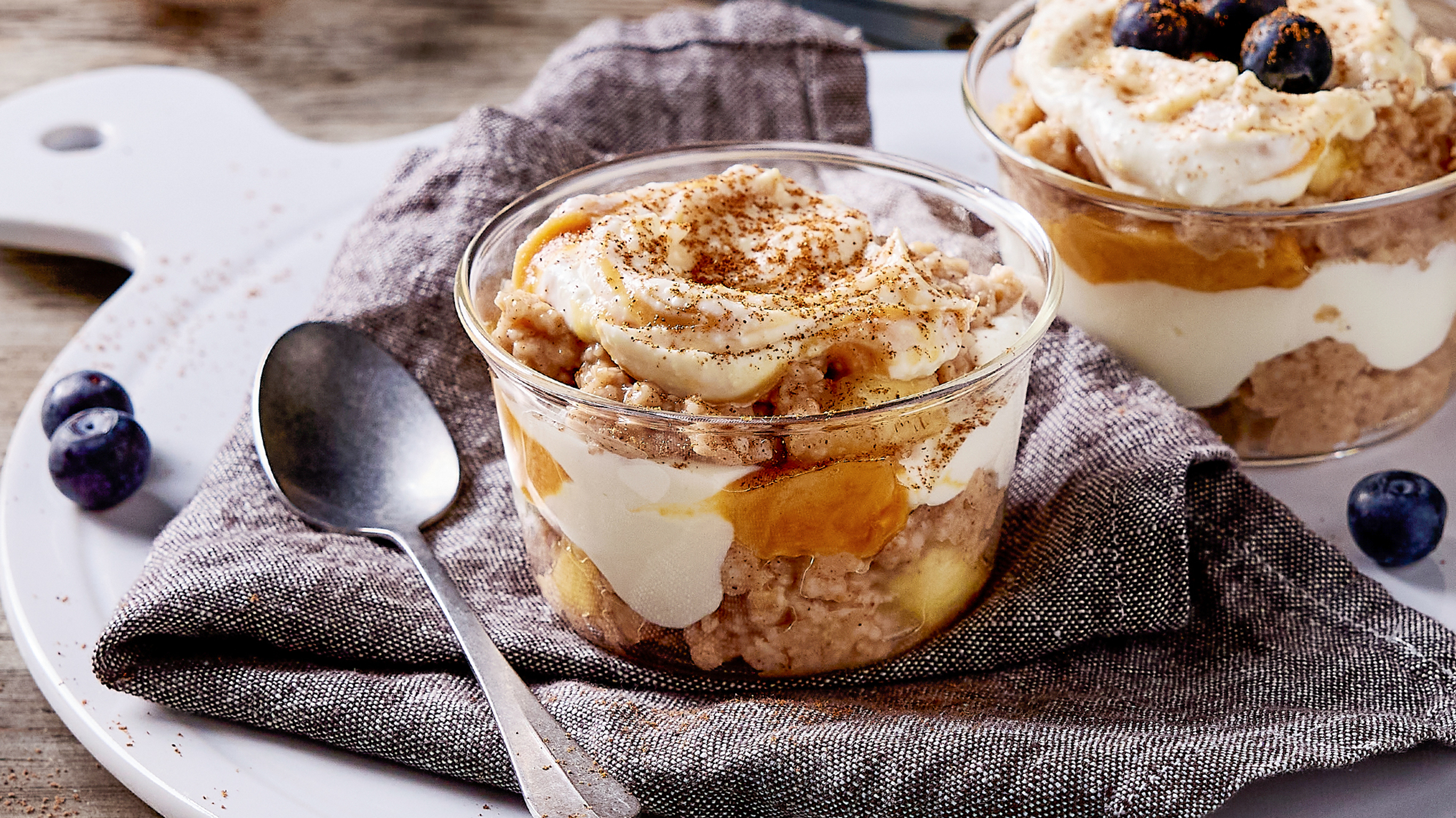 Two glass pots of Cent's Swirled banana & oat pots are decorated with blueberries and ready for eating.