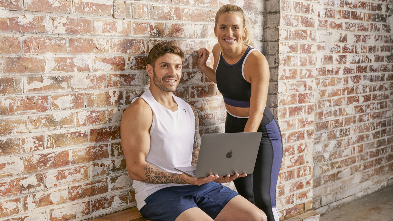 Centr trainers Luke Zocchi and Alexz Parvi look up from a laptop resting on Luke's to smile at the camera.