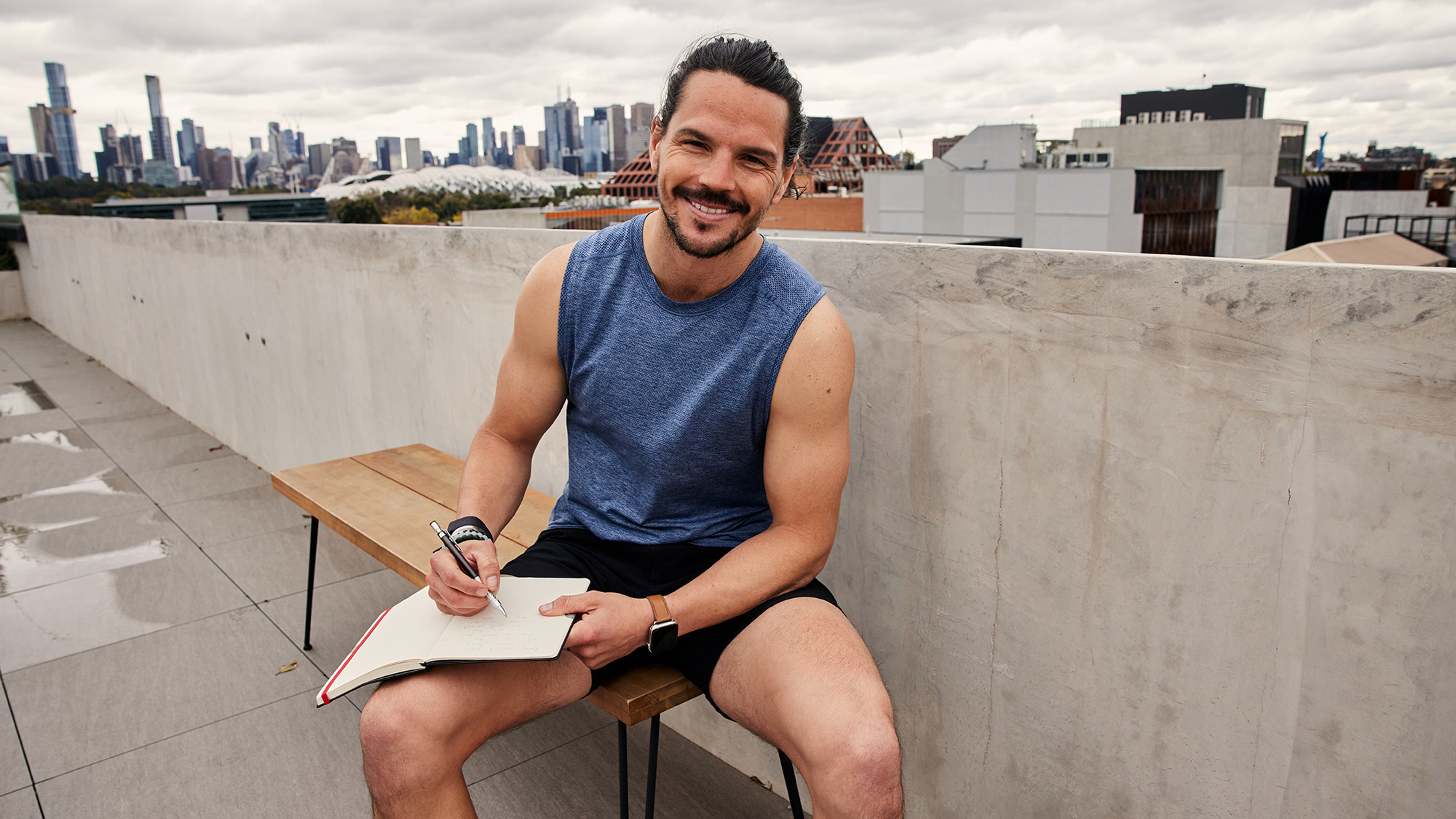 Centr trainer sits on a bench on a city rooftop, journal open on his lap