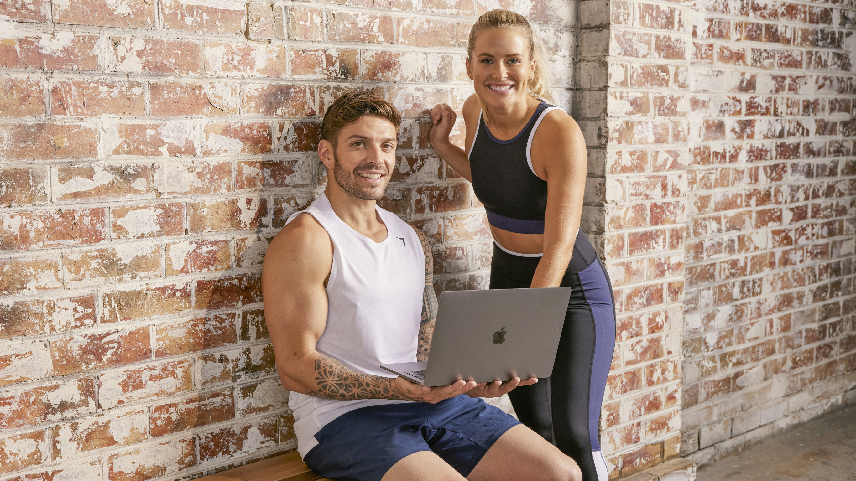 Centr trainers Luke Zocchi and Alexz Parvi look up from a laptop resting on Luke's to smile at the camera.