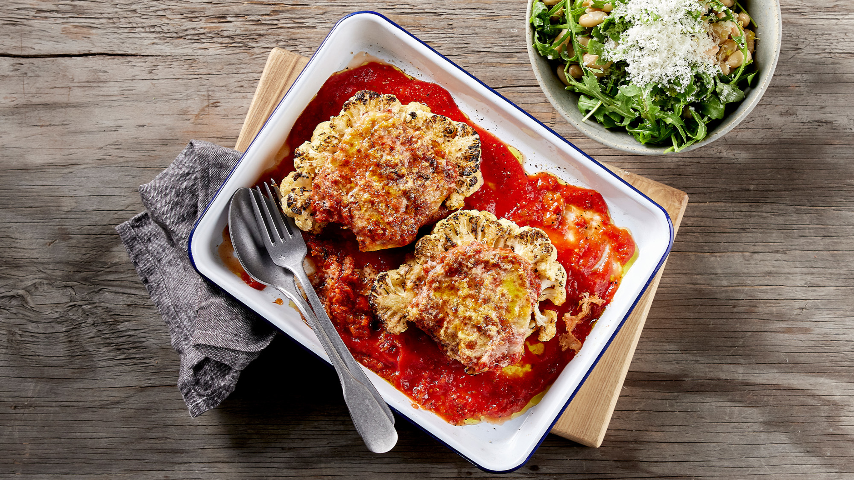 An enamel baking tray of Centr's Six-ingredient Roasted Cauliflower Parmesan sits on wooden board, ready for serving.