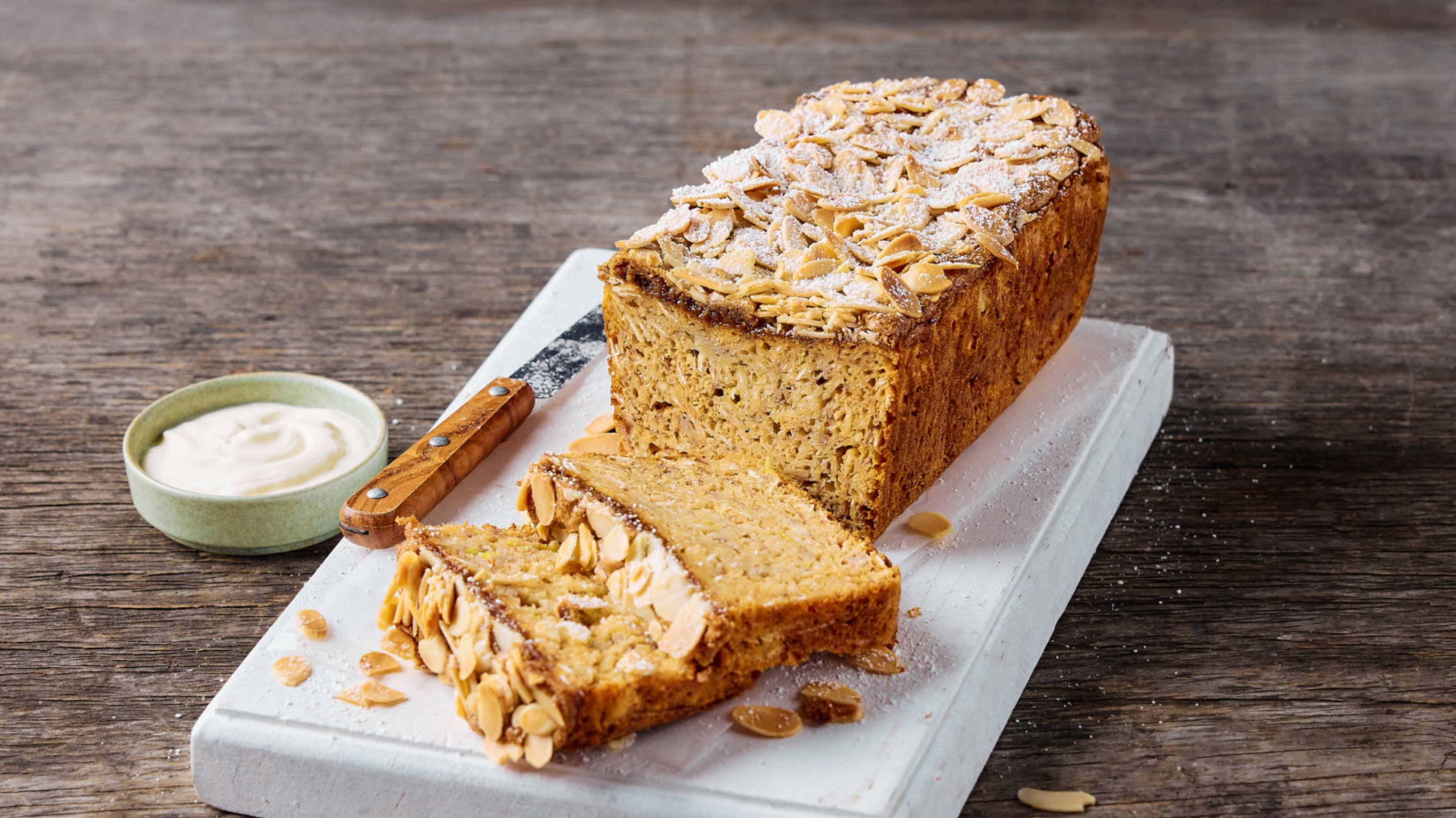 A loaf of Centr's Almond Croissant Banana Bread sits on a cutting board, with a slice cut and ready for eating.