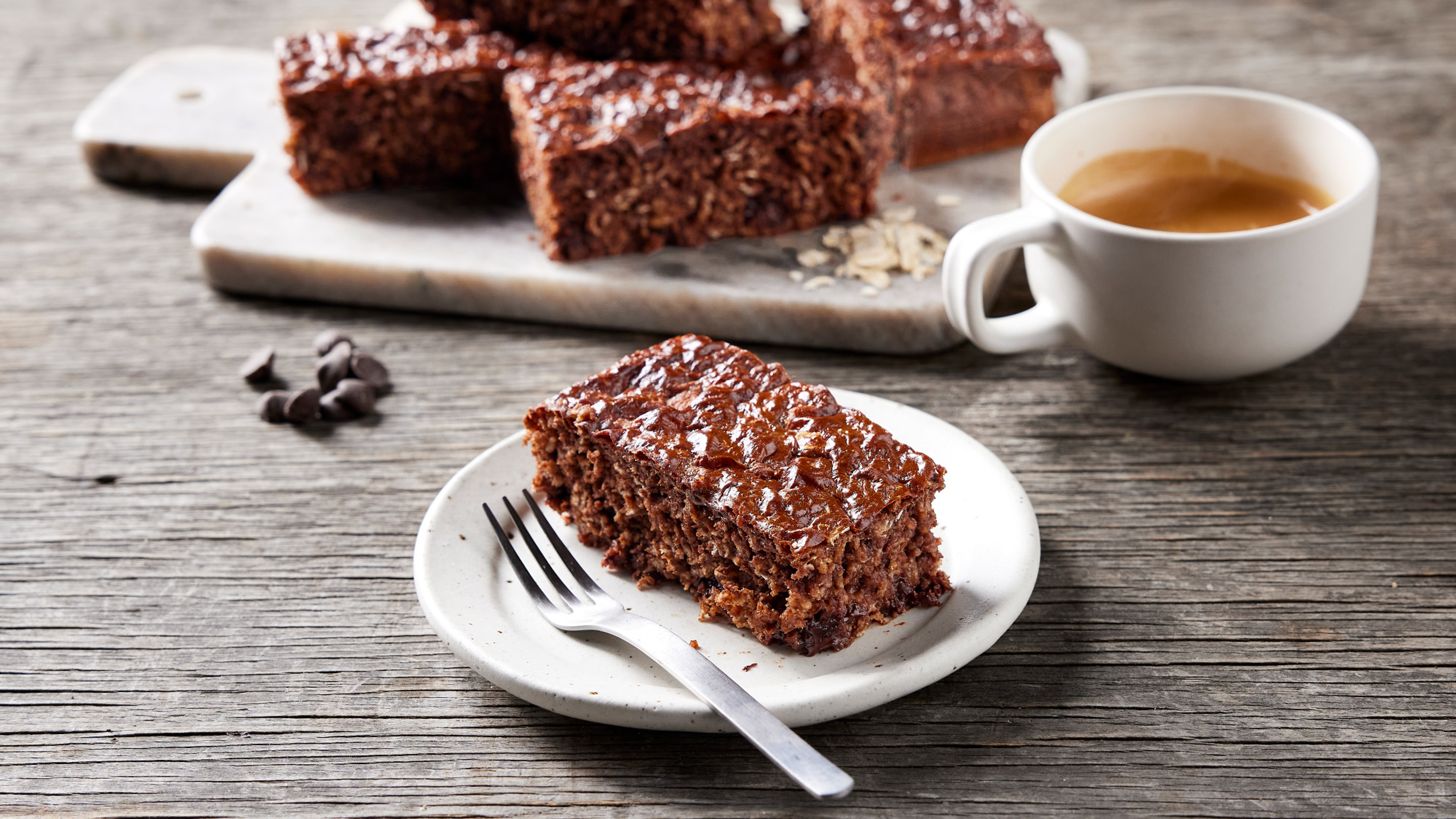 A slice of Chocolate Brownie Oat Slice from the Centr menu sits on a small white plate, with a sliver fork on the side, ready for eating.