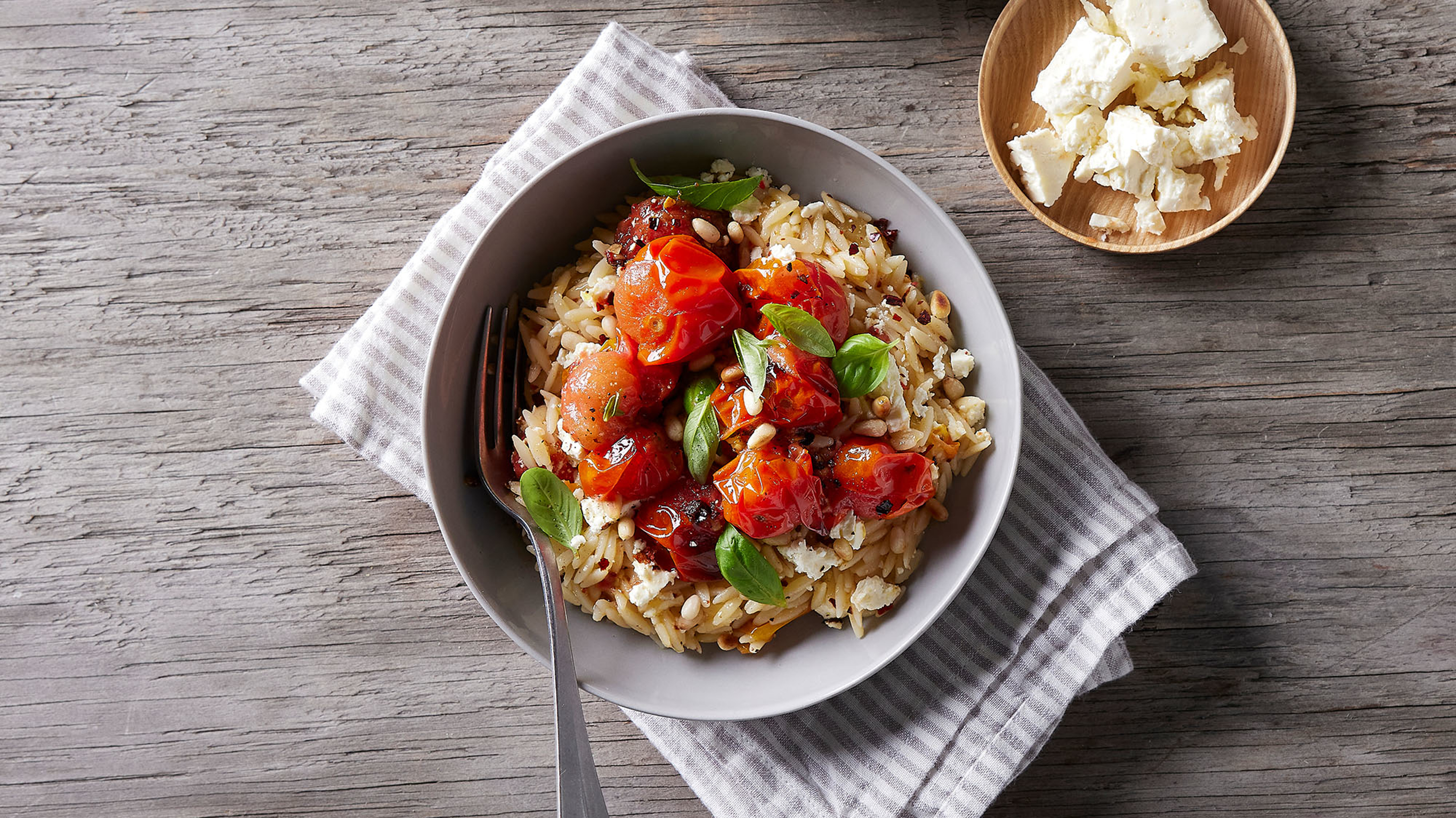 A bowl of Centr's Roasted Tomato Salad with Risoni & Feta sits on a folded grey-striped napkin, ready for eating.