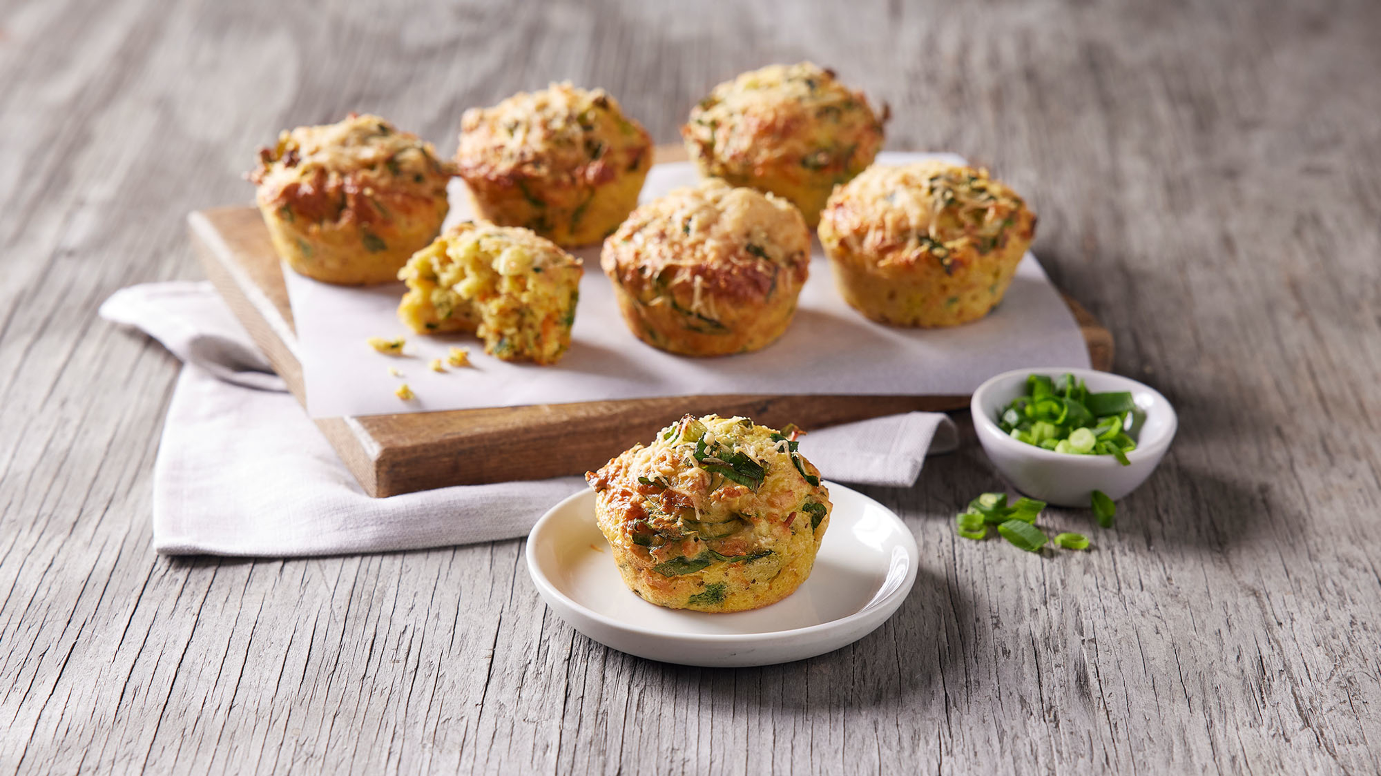 A Spinach, Cheese & Carrot Breakfast Muffin sits on a small plate in front of a wooden cutting board with more muffins ready for eating.