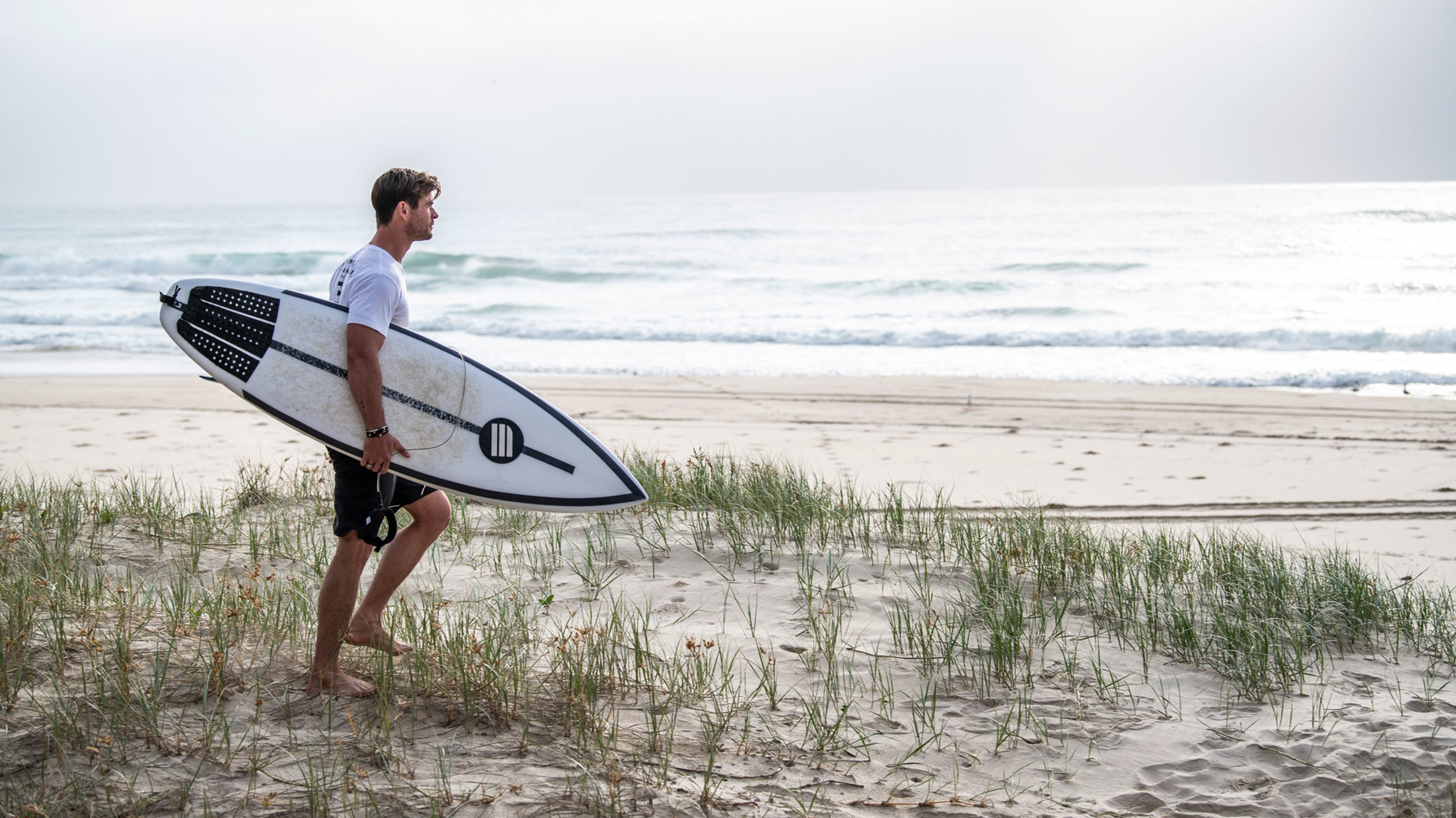Chris Hemsworth on the beach with a surfboard.