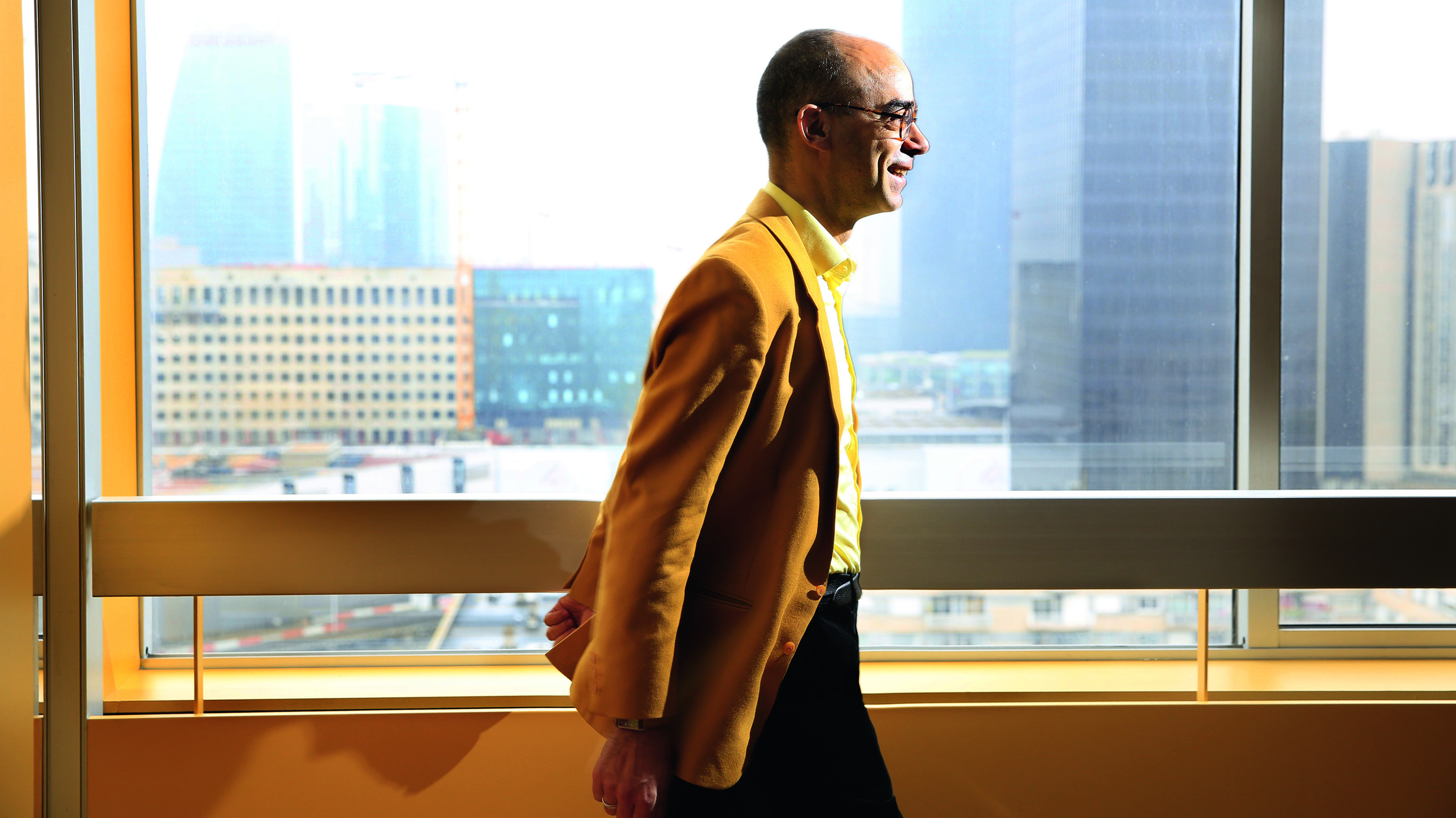 Fabrice Midal walking down a sun lit hallway wearing a mustard-colored jacket.