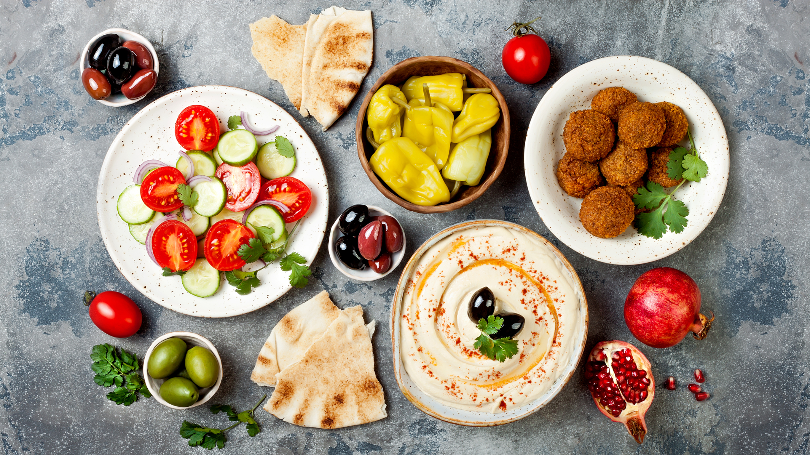 Tomato and cucumber salad, pita bread, hummus and falafal on a grey counter.