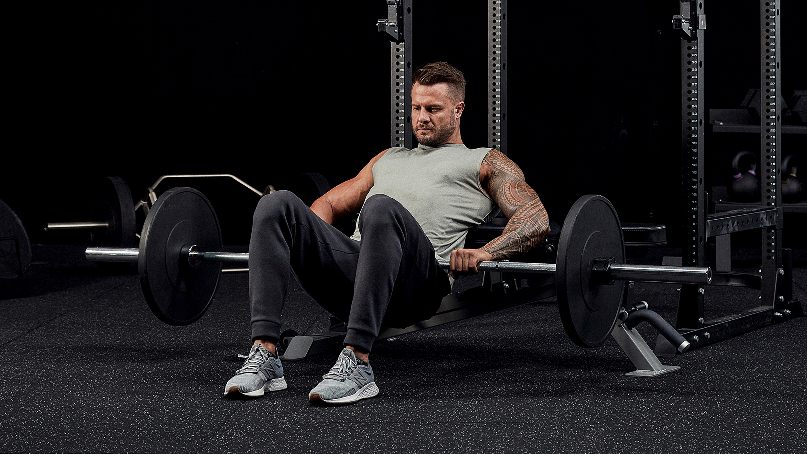 Bobby Holland Hanton prepares to perform a glute thrust using a bar and weight plates in gym.