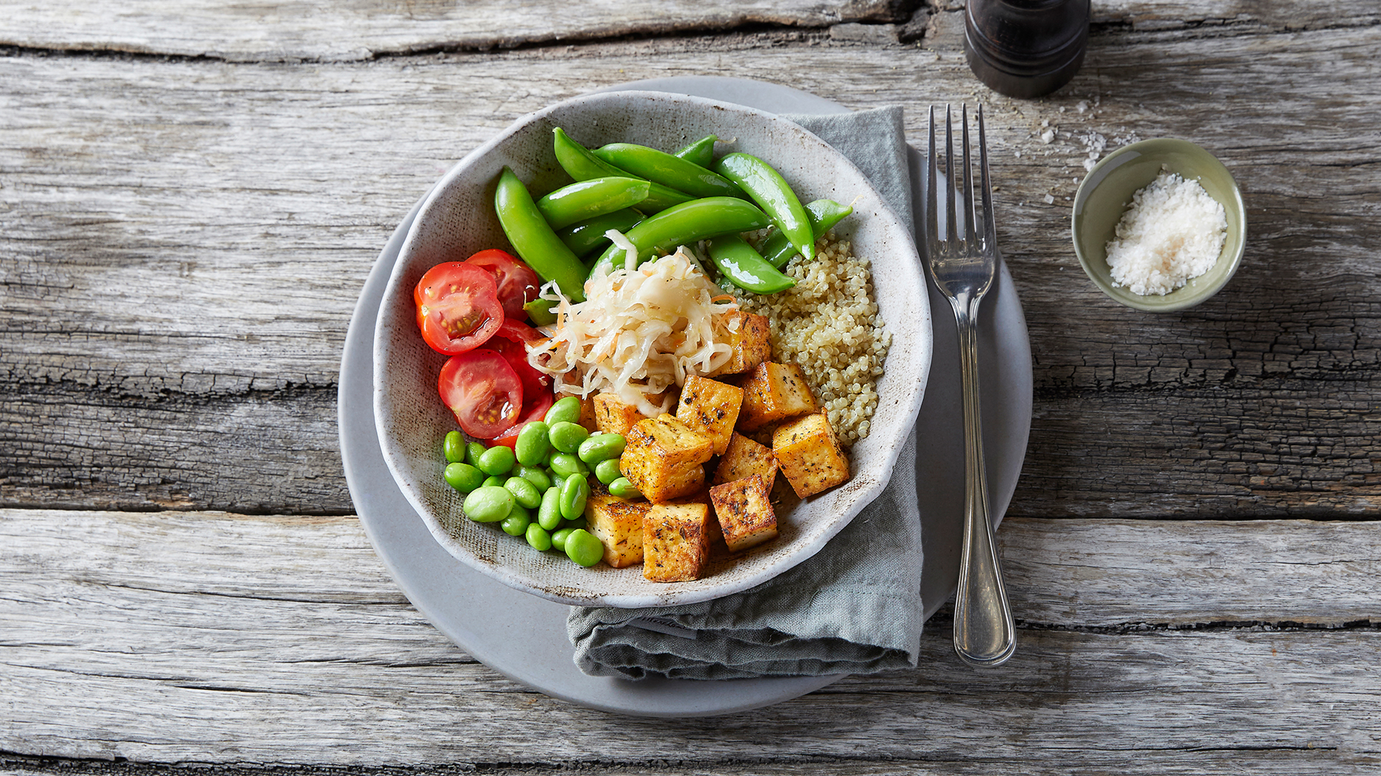 A bowl of Centr's Buddha Bowl with Sauerkraut sits on a matching plate with napkin and fork on a wooden table.