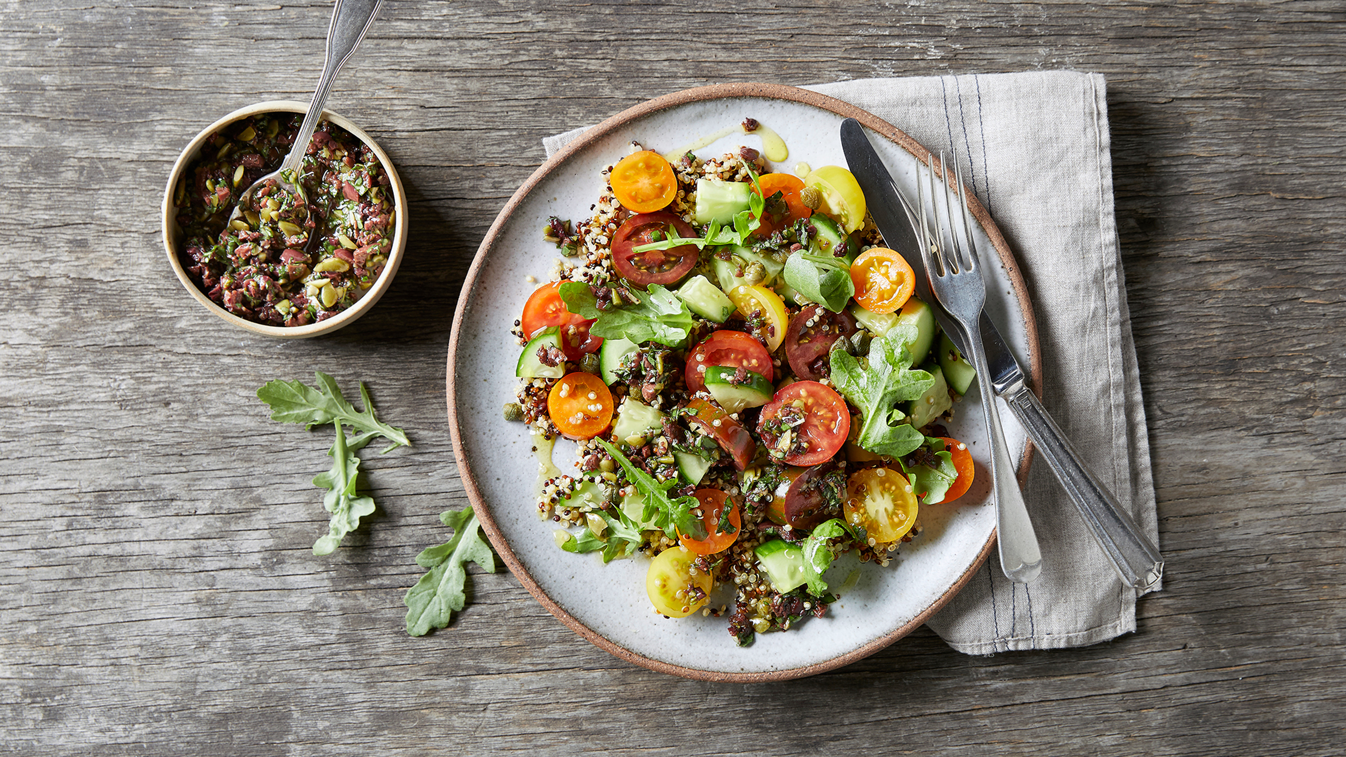 A bowl of Centr's Mediterranean Quinoa Salad sits on a wooden table with a side dish of chunky olive tapenade.