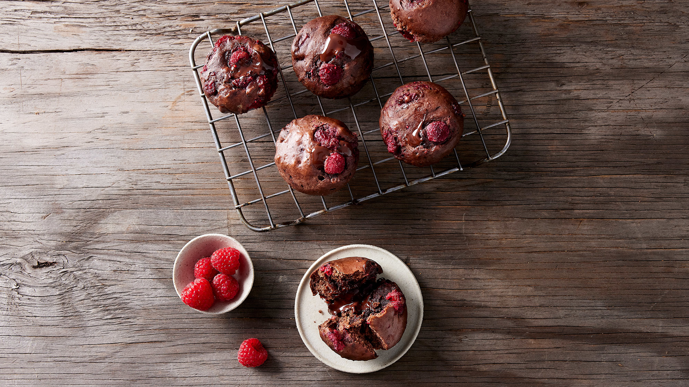 5 of Centr's Gooey Chocolate & Raspberry Muffins rest on a cooling rack, and another sits quartered on a plate ready for eating.